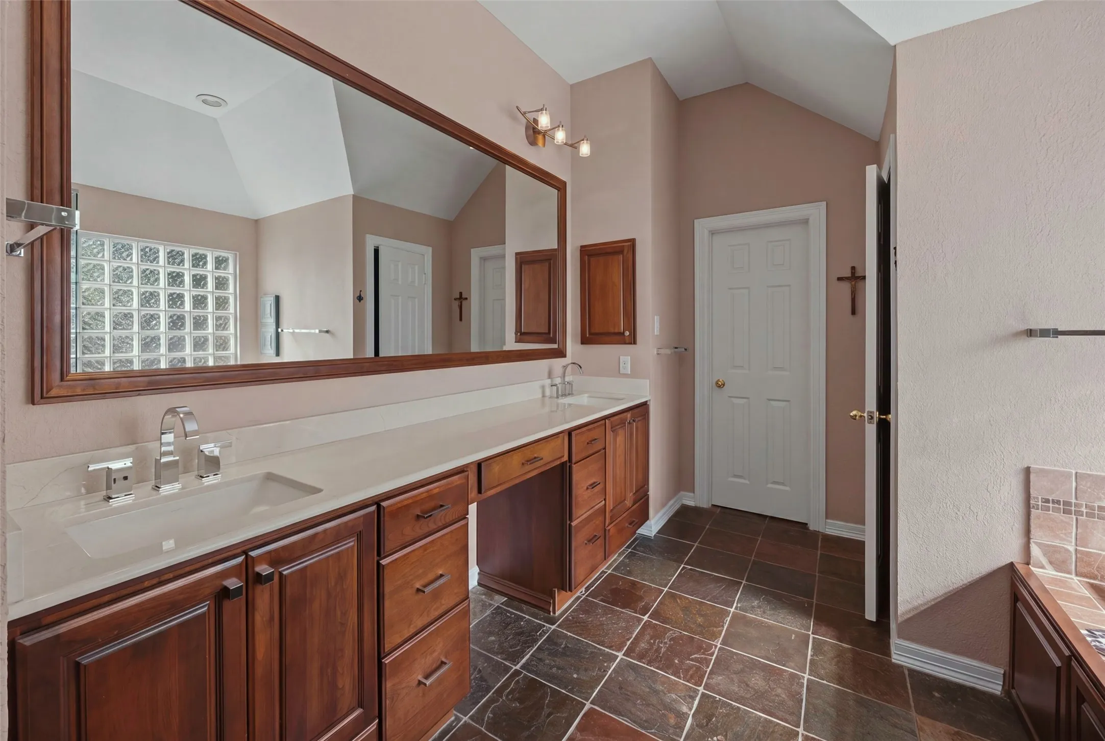 Vaulted ceiling, dual vanity with undermount sinks, wood-finish cabinetry, stone tile flooring, and a glass block window feature