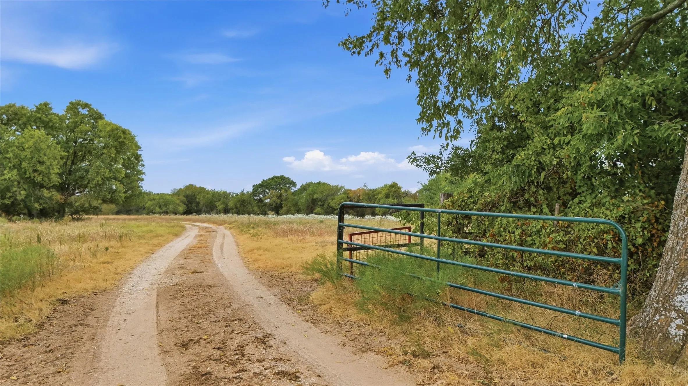 Gated entry to a private unpaved road, surrounded by mature trees and open fields