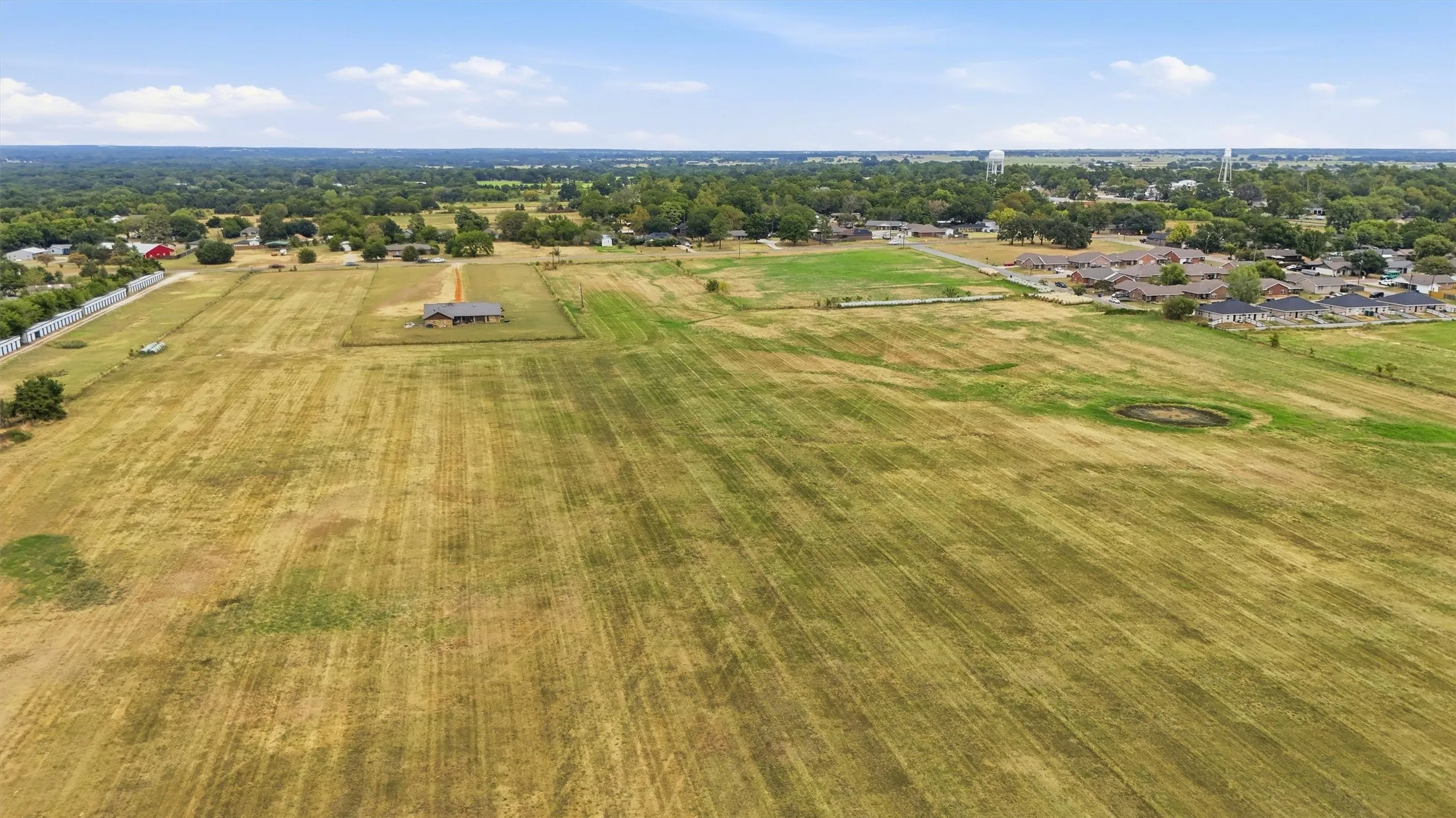 Expansive open land with a residential structure, scattered trees, and distant community features