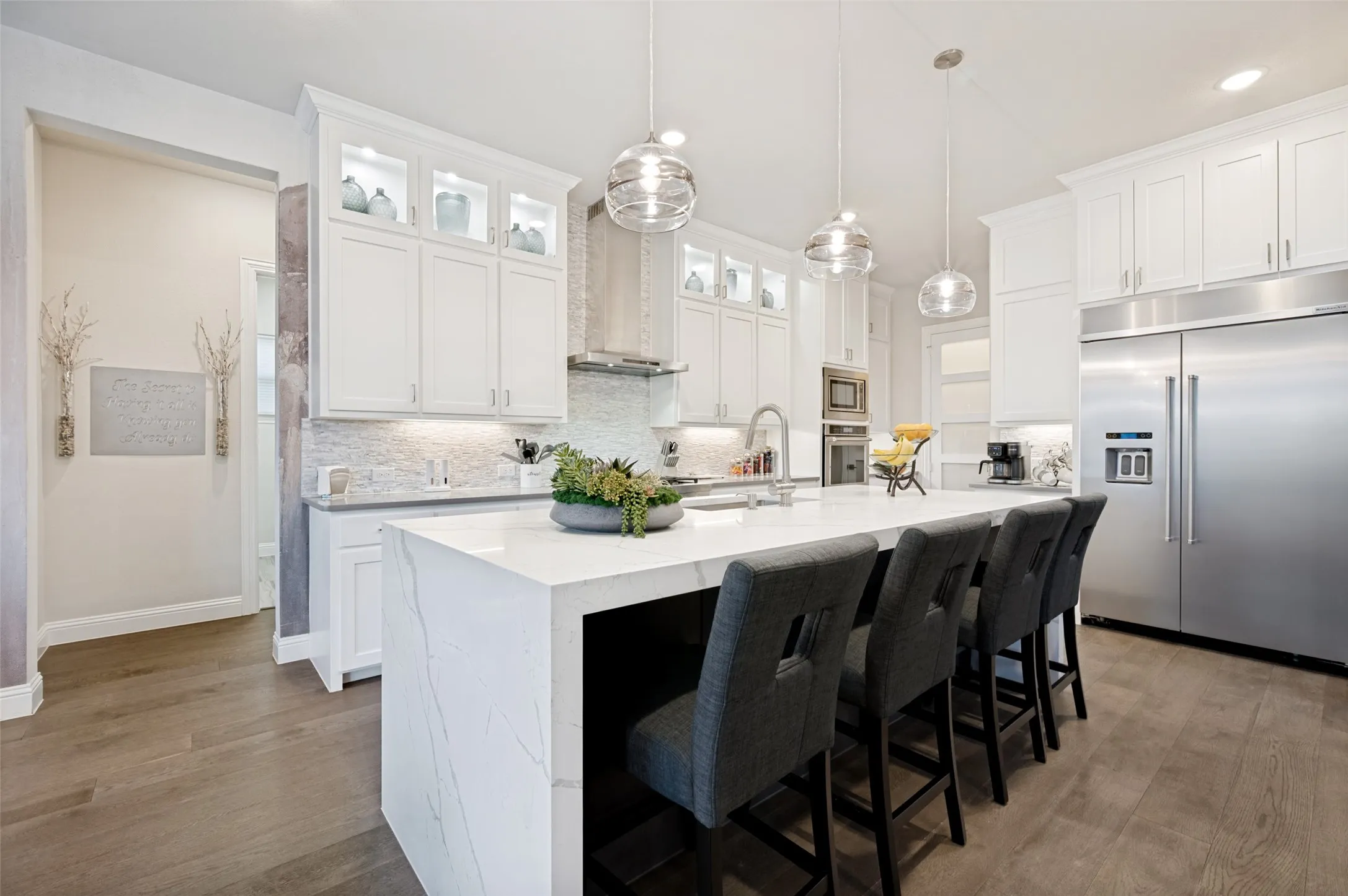 Spacious kitchen featuring an expansive island with a waterfall edge, stainless steel appliances, white cabinetry with display lighting, and wood-finish flooring