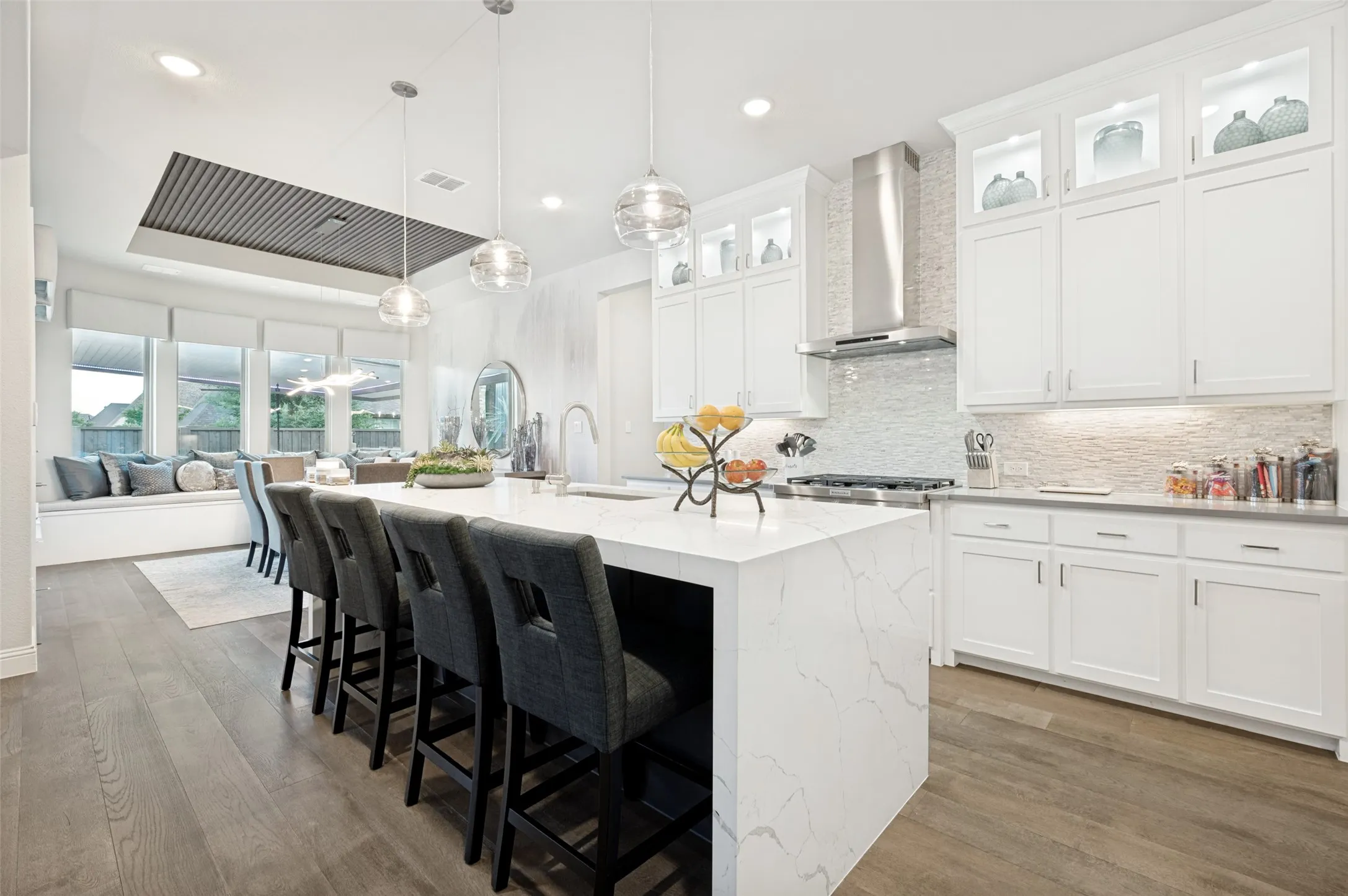 Waterfall-edge island with seating, wood-finish flooring, white cabinetry with display lighting, built-in banquette seating, and a stainless steel range hood