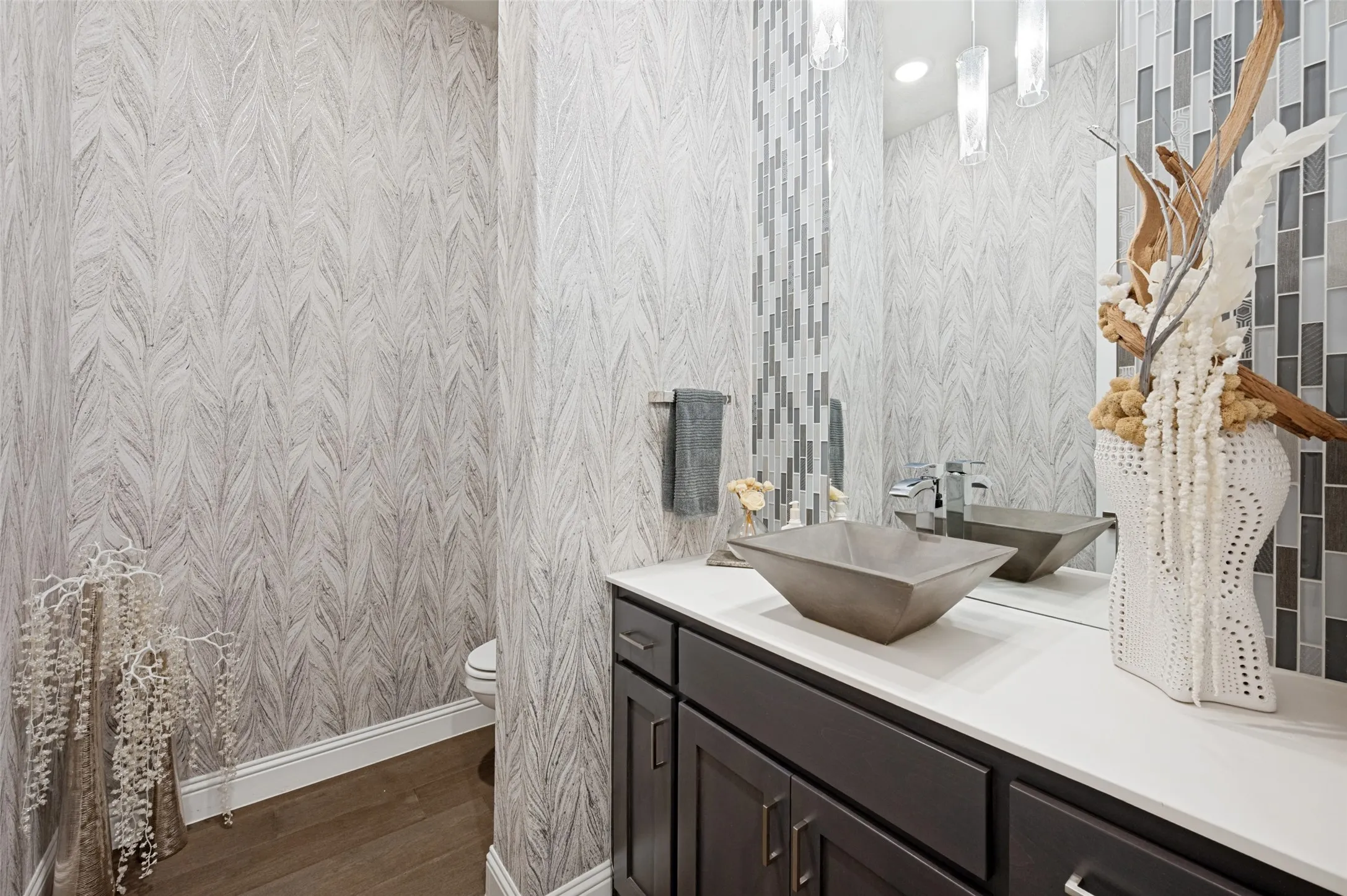 Powder room with patterned wall covering, a dark wood-finish vanity, white countertop, square vessel sink, and rectangular tile backsplash