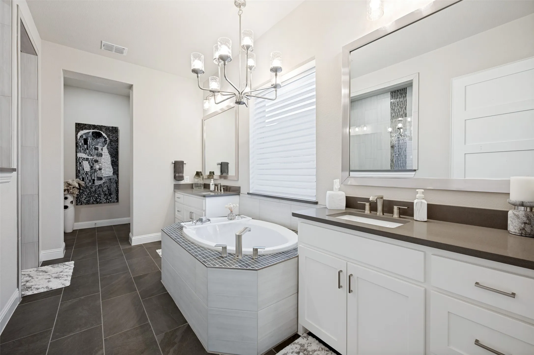 Floor-set bathtub with mosaic tile surround, dual vanities with white cabinetry, brown countertops, and large framed mirrors