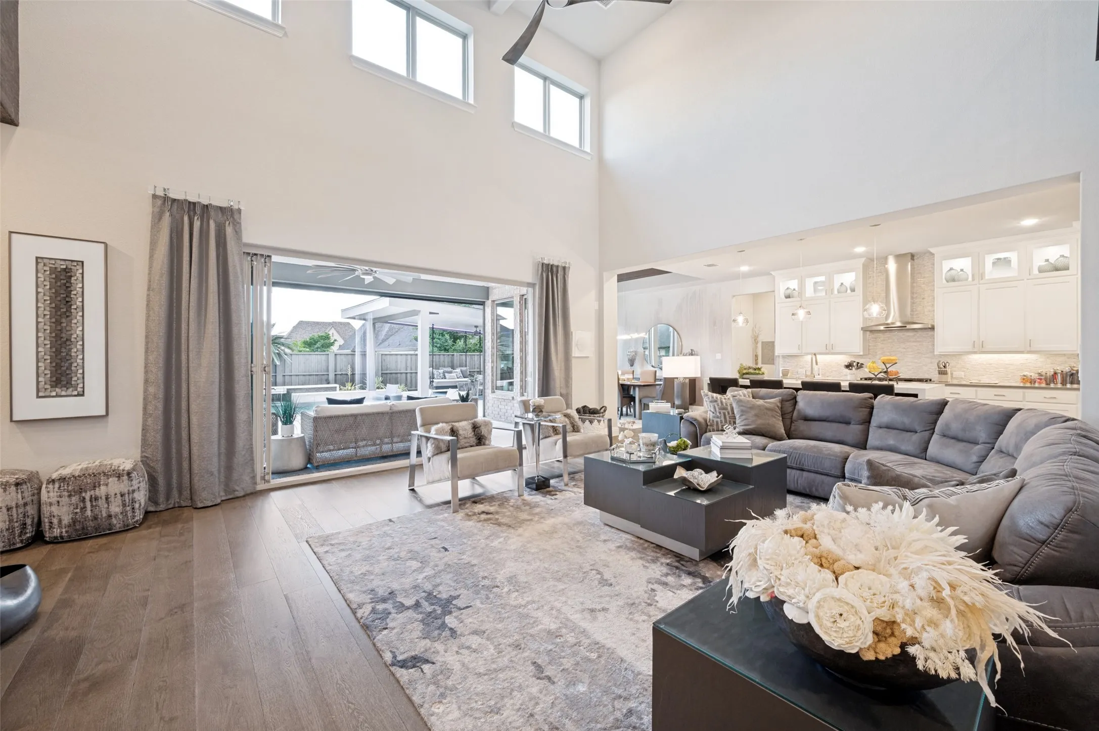 Open-concept living area featuring a two-story ceiling with transom windows, wood-finish flooring, and a large sliding glass door opening to an outdoor patio with a pool