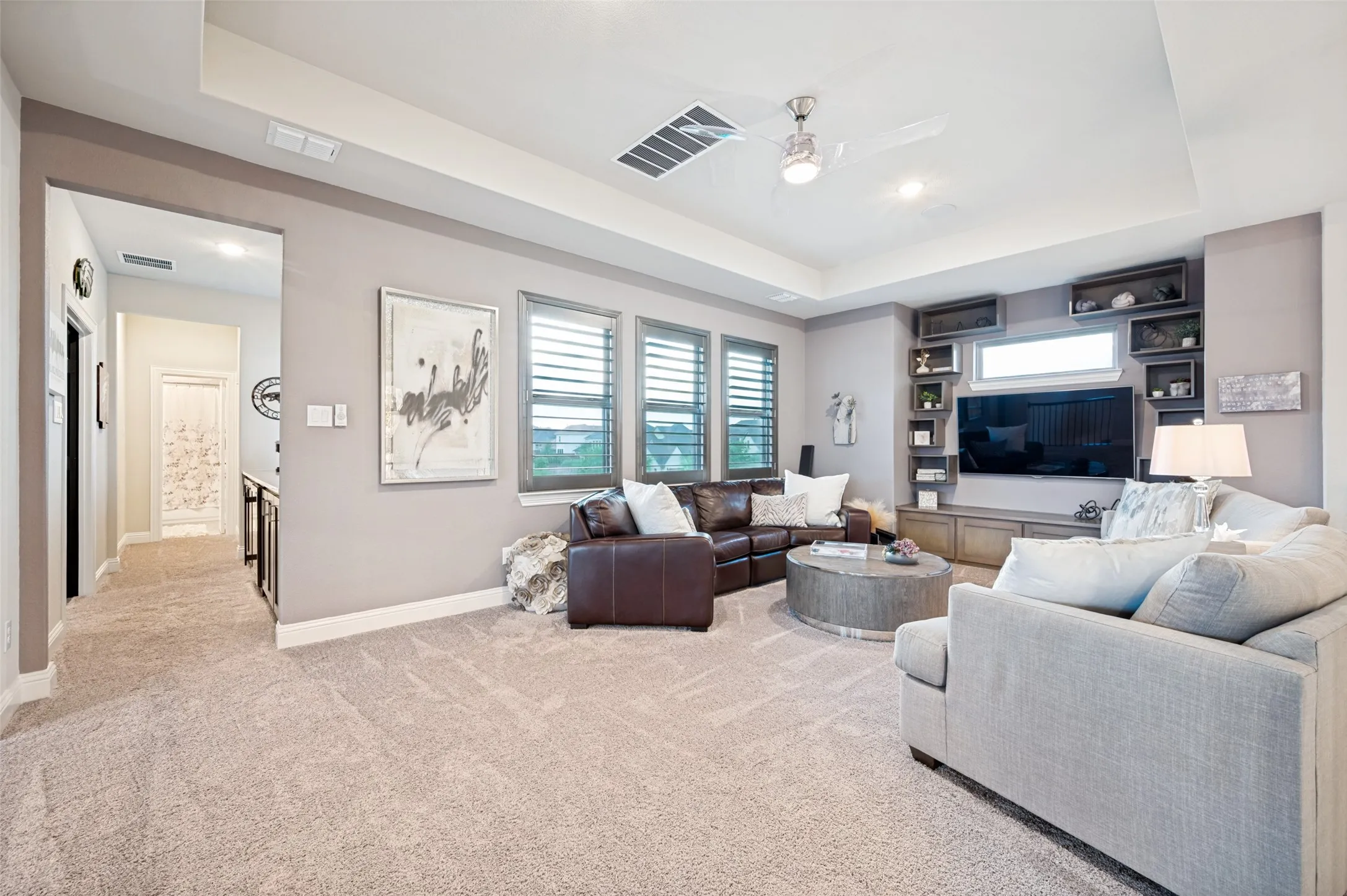 Spacious living area featuring neutral carpeting, a tray ceiling with a ceiling fan, and multiple windows with plantation shutters