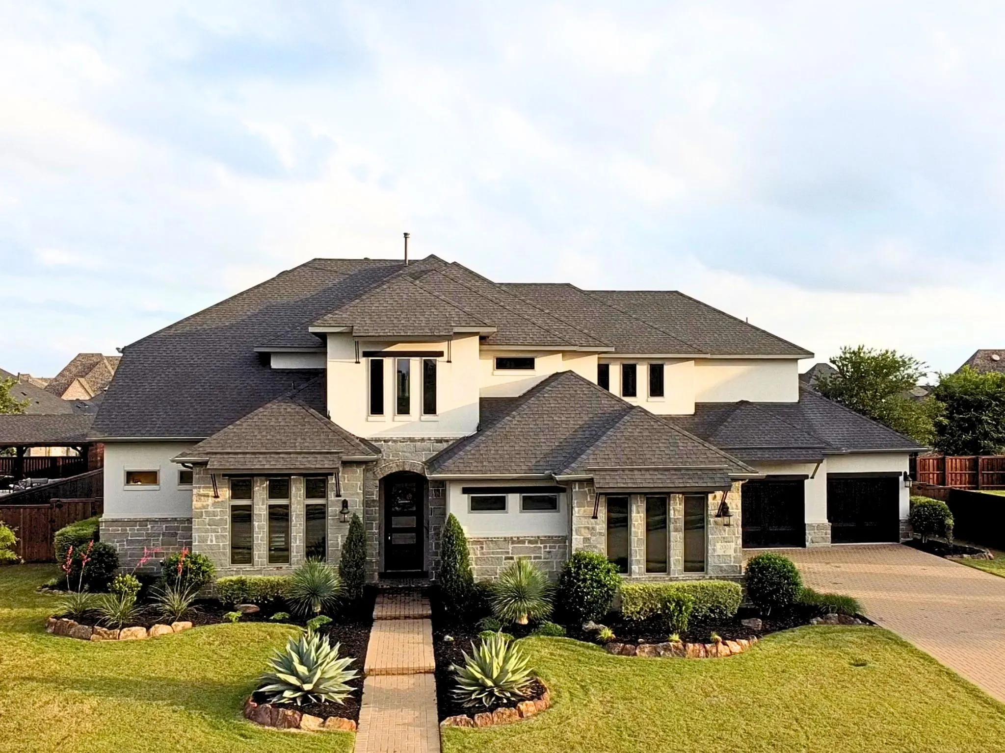 Stucco and stone exterior, dark shingle roof, integrated two-car garage, brick paver walkway, and structured landscaping