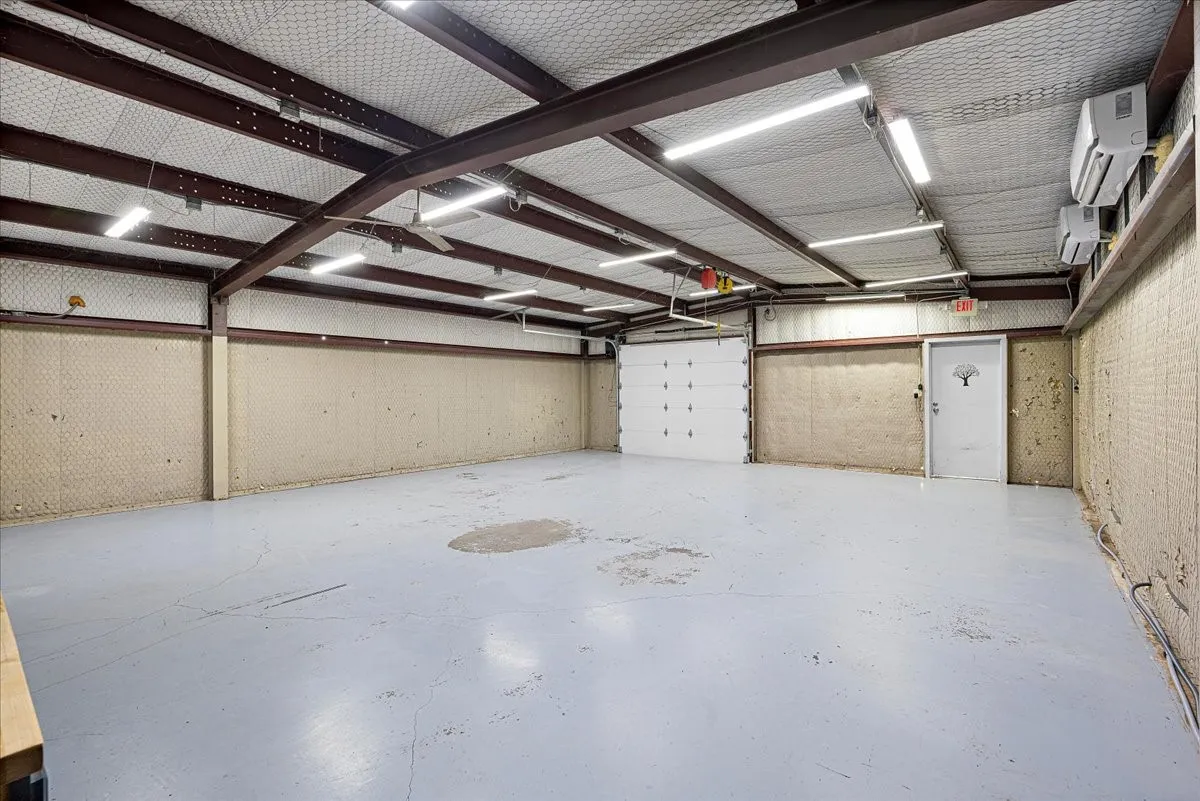 Expansive open space featuring high ceilings with exposed metal beams, multiple fluorescent light fixtures, a large roll-up door, a standard entry door, and multiple wall-mounted air conditioning units