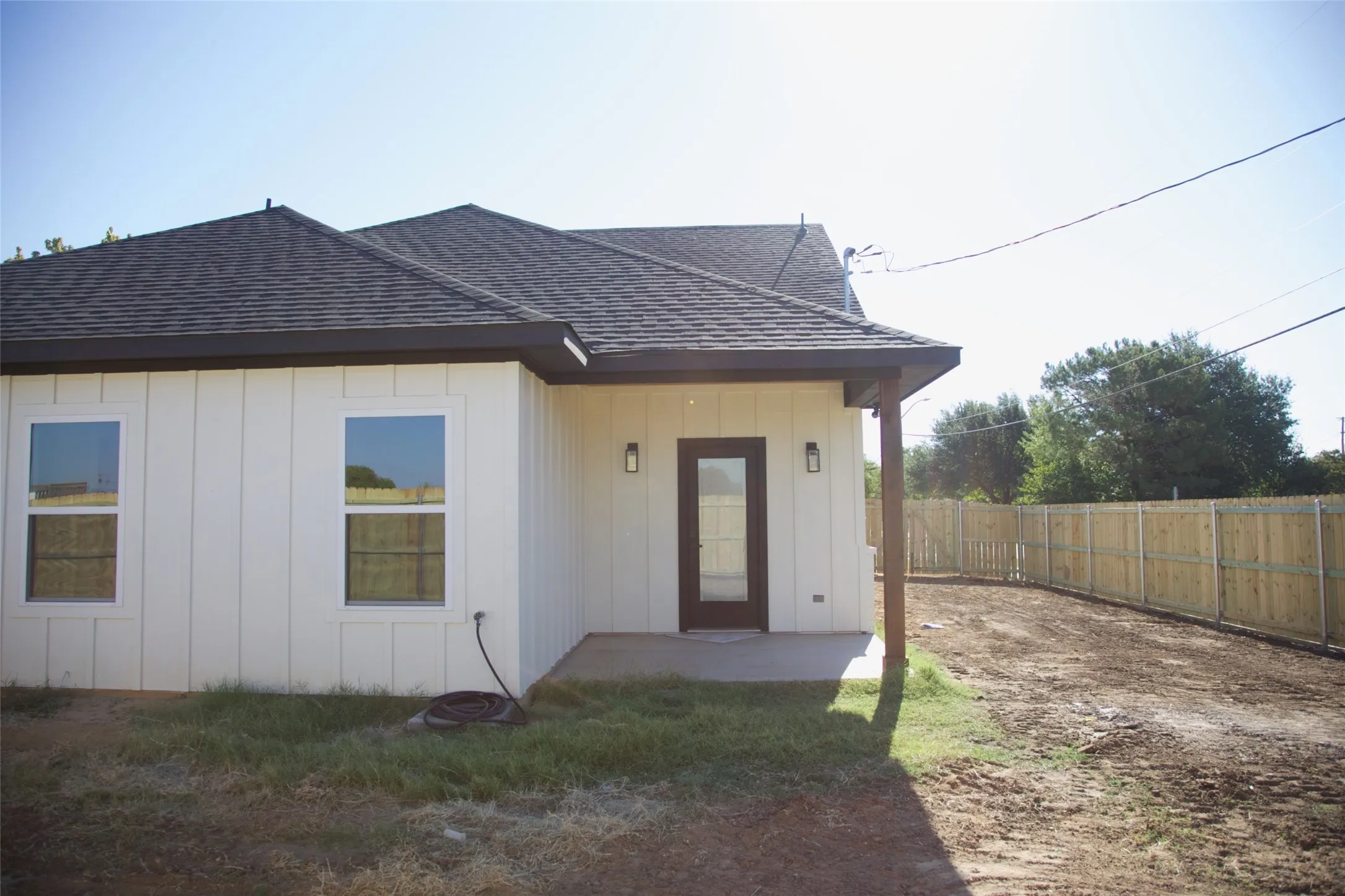 Board and batten siding, dark-shingle roof, covered patio with support column, fenced yard, and exterior lighting fixtures