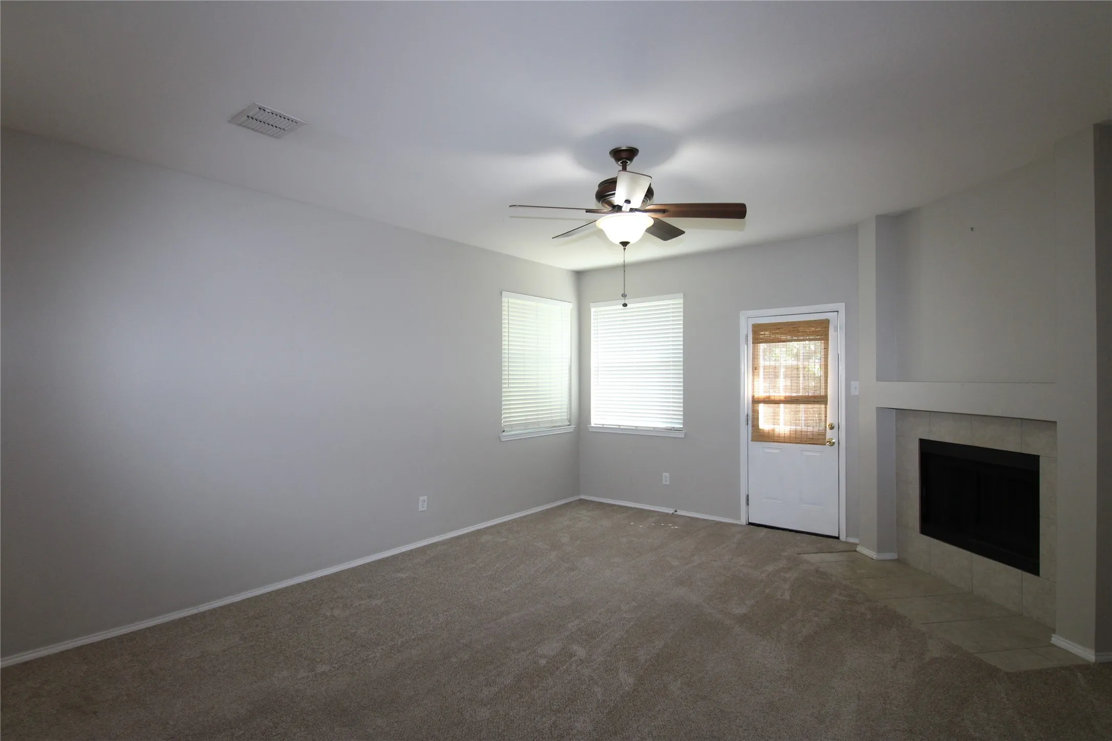 Carpeted living area featuring a ceiling fan with integrated lighting, two windows with blinds, a white door, and a tiled fireplace with a mantel