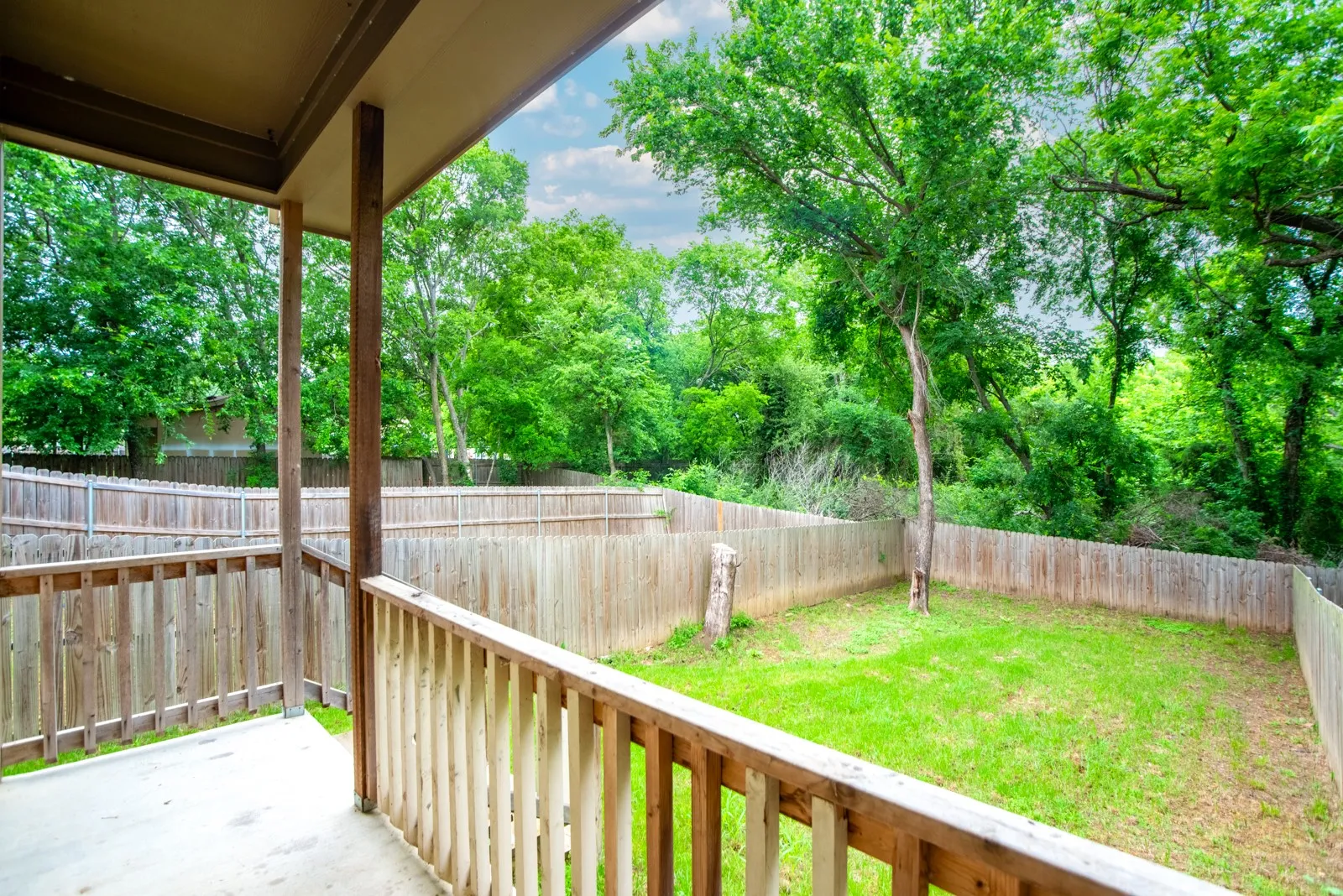 Covered patio with a concrete surface and wooden railing, overlooking a fenced backyard with a grass lawn and mature trees