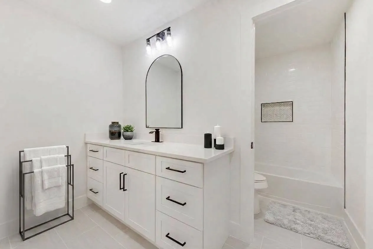 White vanity with quartz countertop, black hardware, arched mirror, and tiled shower with built-in niche