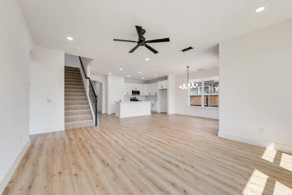 Light wood-finish flooring throughout, modern kitchen with white cabinetry and stainless steel appliances, contemporary chandelier, recessed lighting, and a staircase with carpeting and an ornate railing