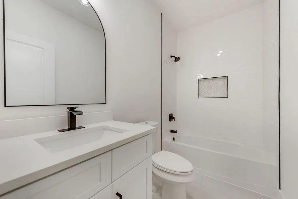 Modern bathroom featuring a white vanity with integrated sink, matte black fixtures, an arched mirror, and a tiled tub surround with an inset niche