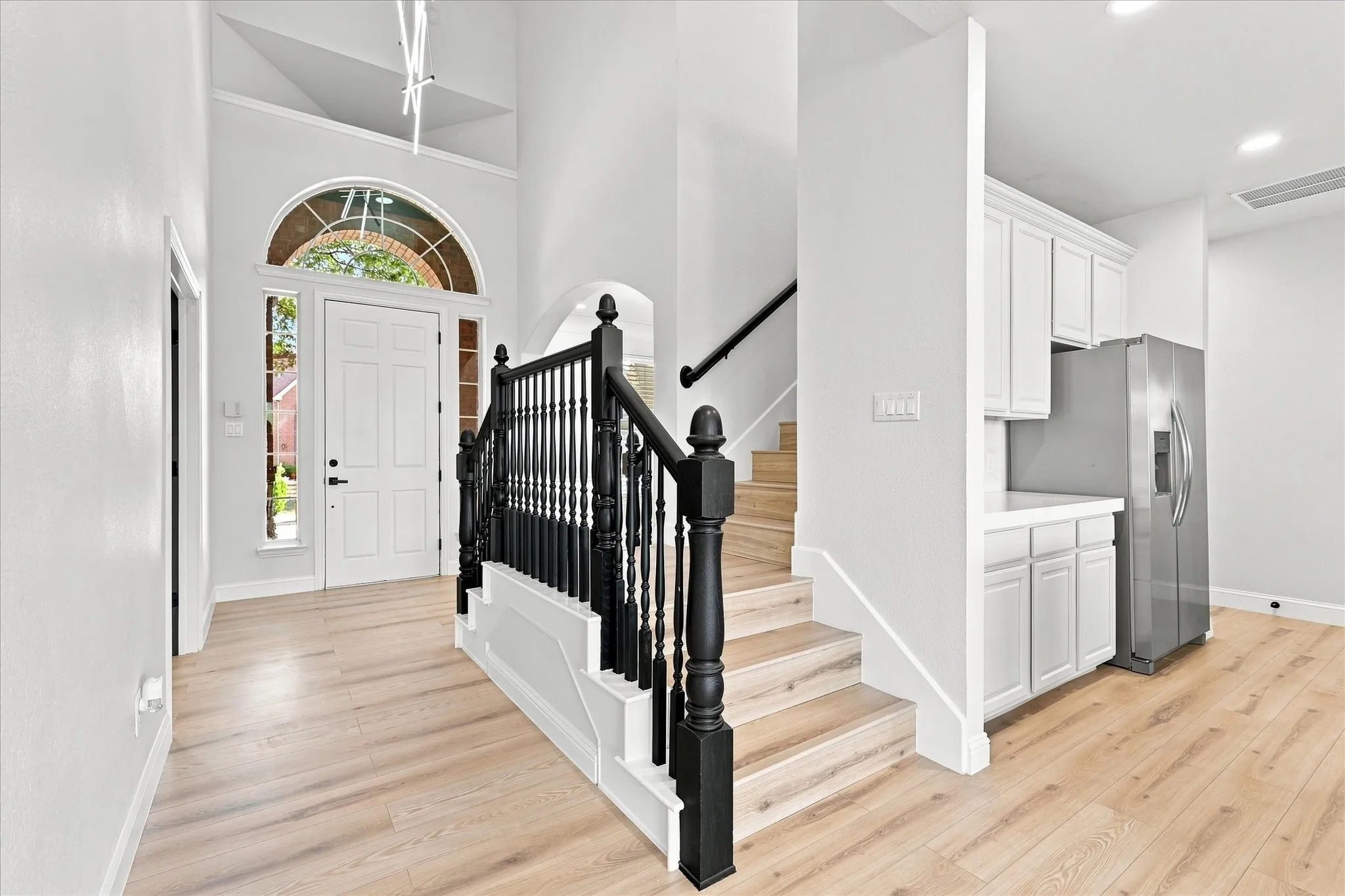 Bright, open foyer featuring a curved staircase with black spindle railings and natural wood treads, arched transom window with sidelights, wide-plank wood-finish flooring throughout, and seamless sightlines to the kitchen featuring white cabinetry, stainless steel appliances, and gray tile backsplash