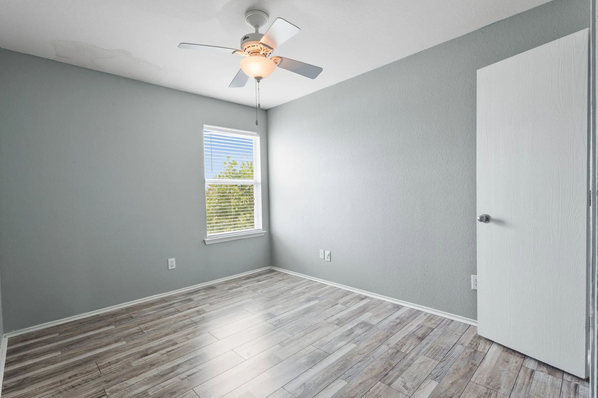 Wood-finish flooring, ceiling fan with integrated lighting, single window with blinds, and a white interior door