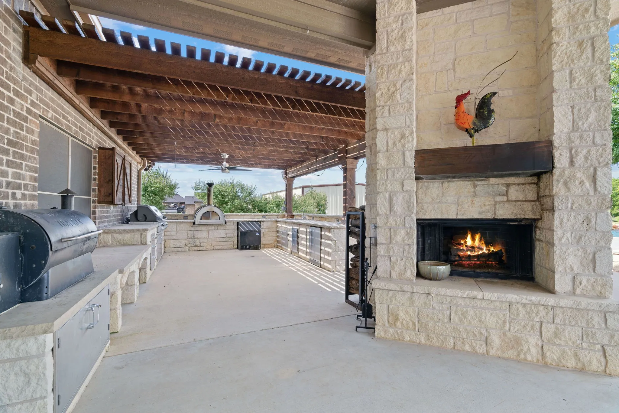 Covered outdoor living space featuring stone-clad fireplace, built-in outdoor kitchen, wood pergola, and ceiling fan