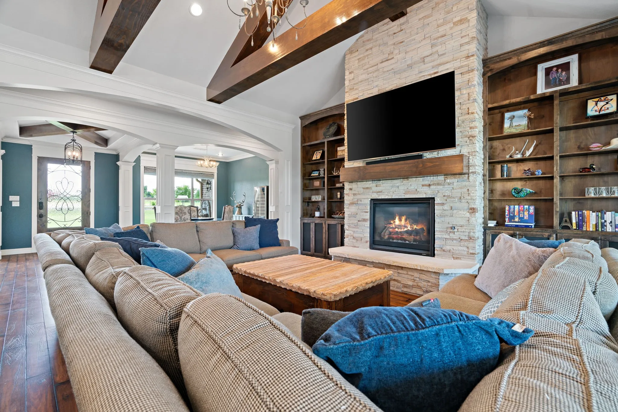 Vaulted ceiling with wood beams, stone-clad fireplace with wood mantel, built-in shelving, wood-finish flooring, and arched doorways