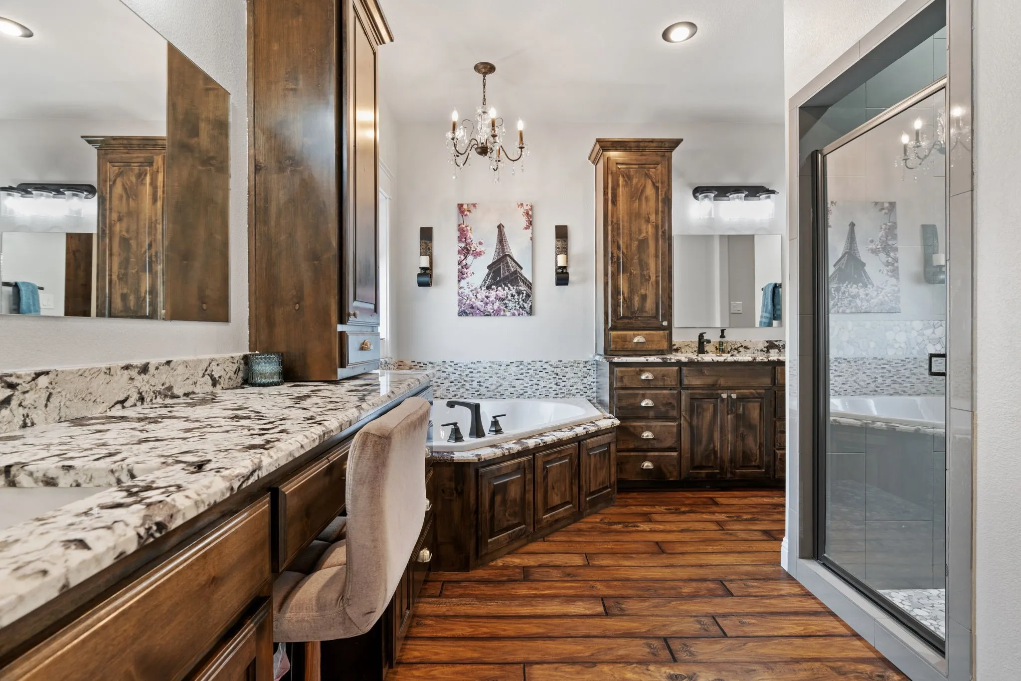 Wood-finish flooring, dual vanities with stone countertops, built-in cabinetry, a corner soaking tub with mosaic tile surround, and a glass-enclosed shower