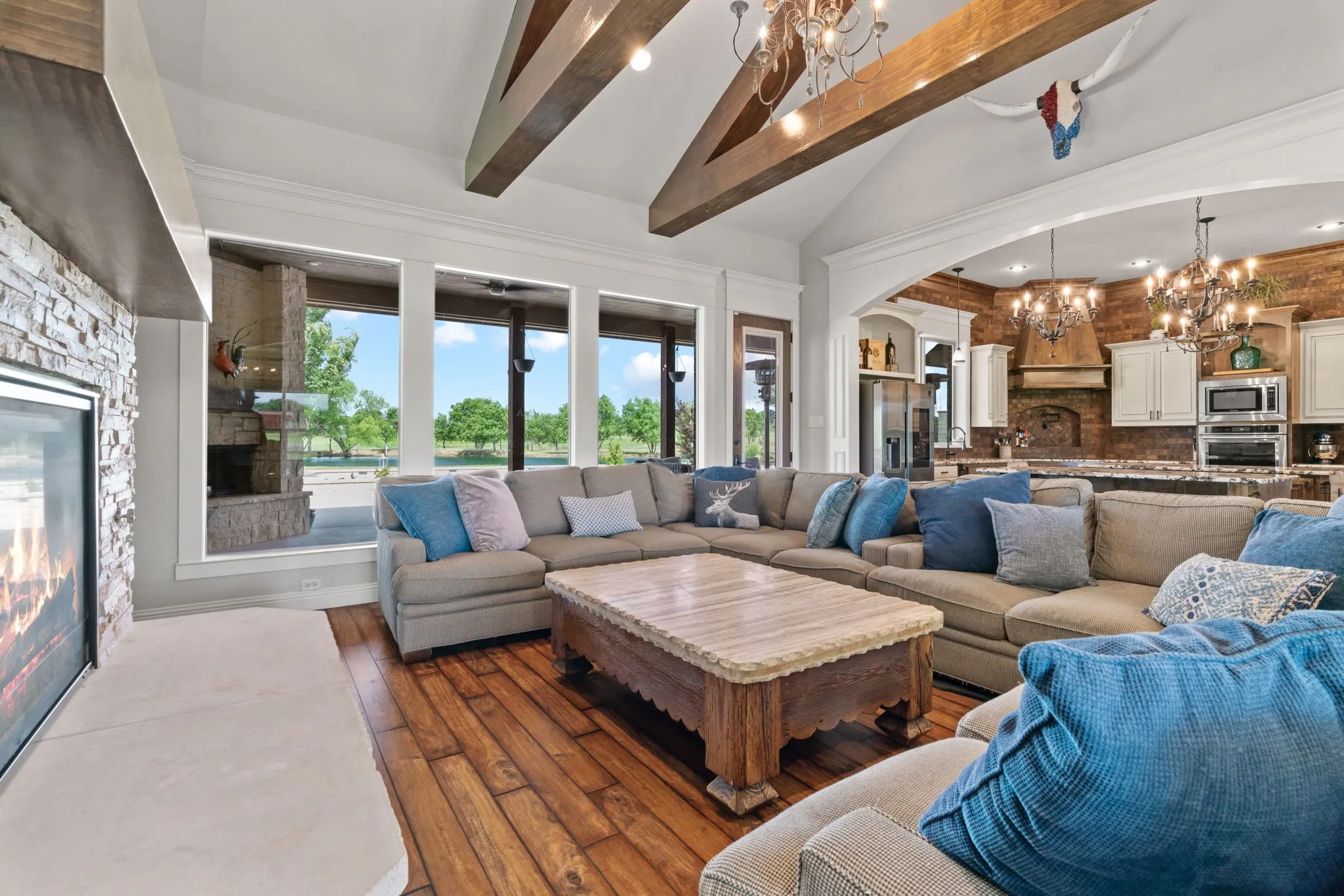 Vaulted ceiling with exposed wood beams, stone-faced fireplace with a hearth, wood-finish flooring, large windows, and a cased opening to the kitchen