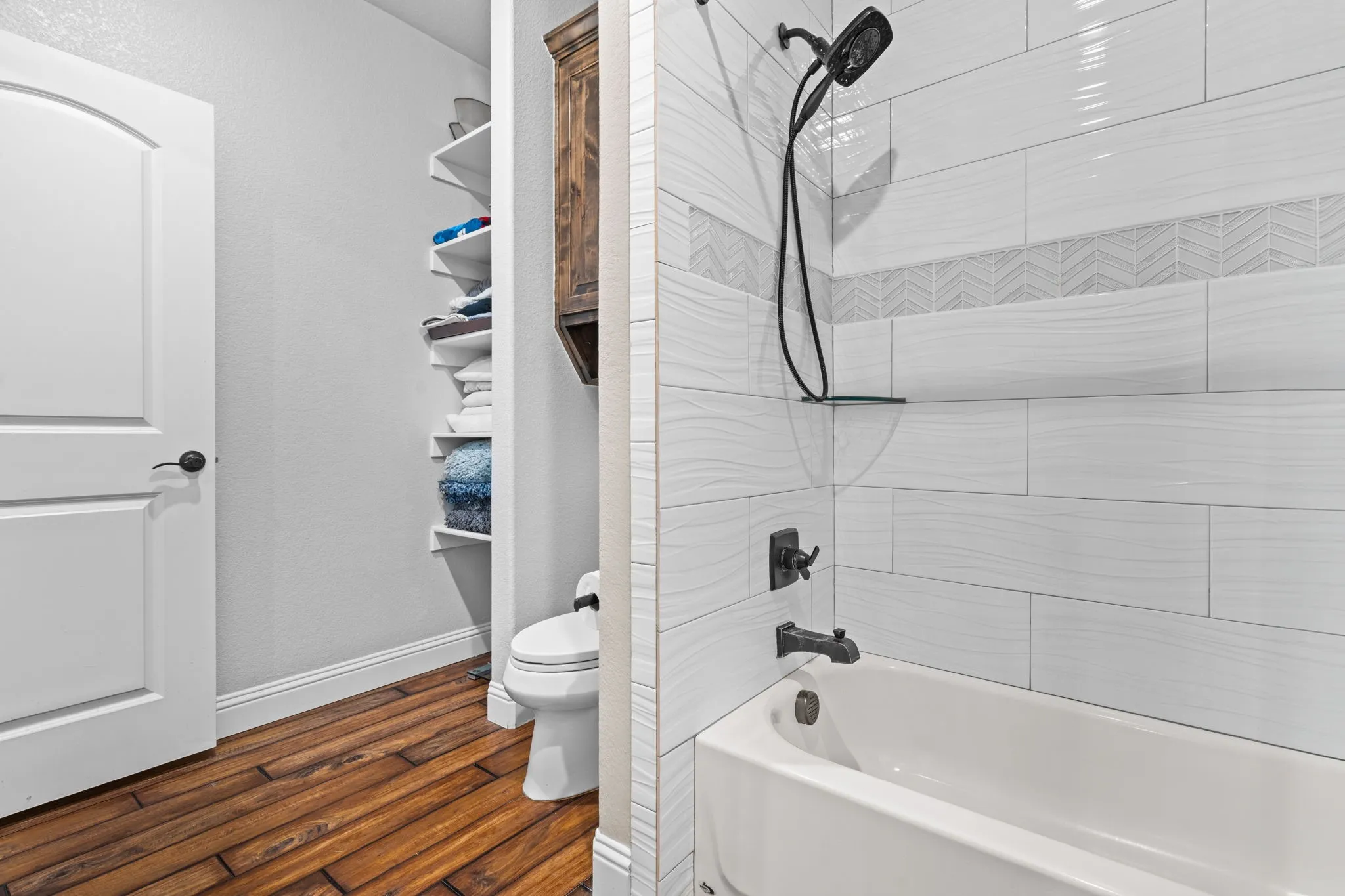 Porcelain tub with matte black fixtures, white subway tile surround with decorative accent band, integrated shelving, and wood-finish flooring