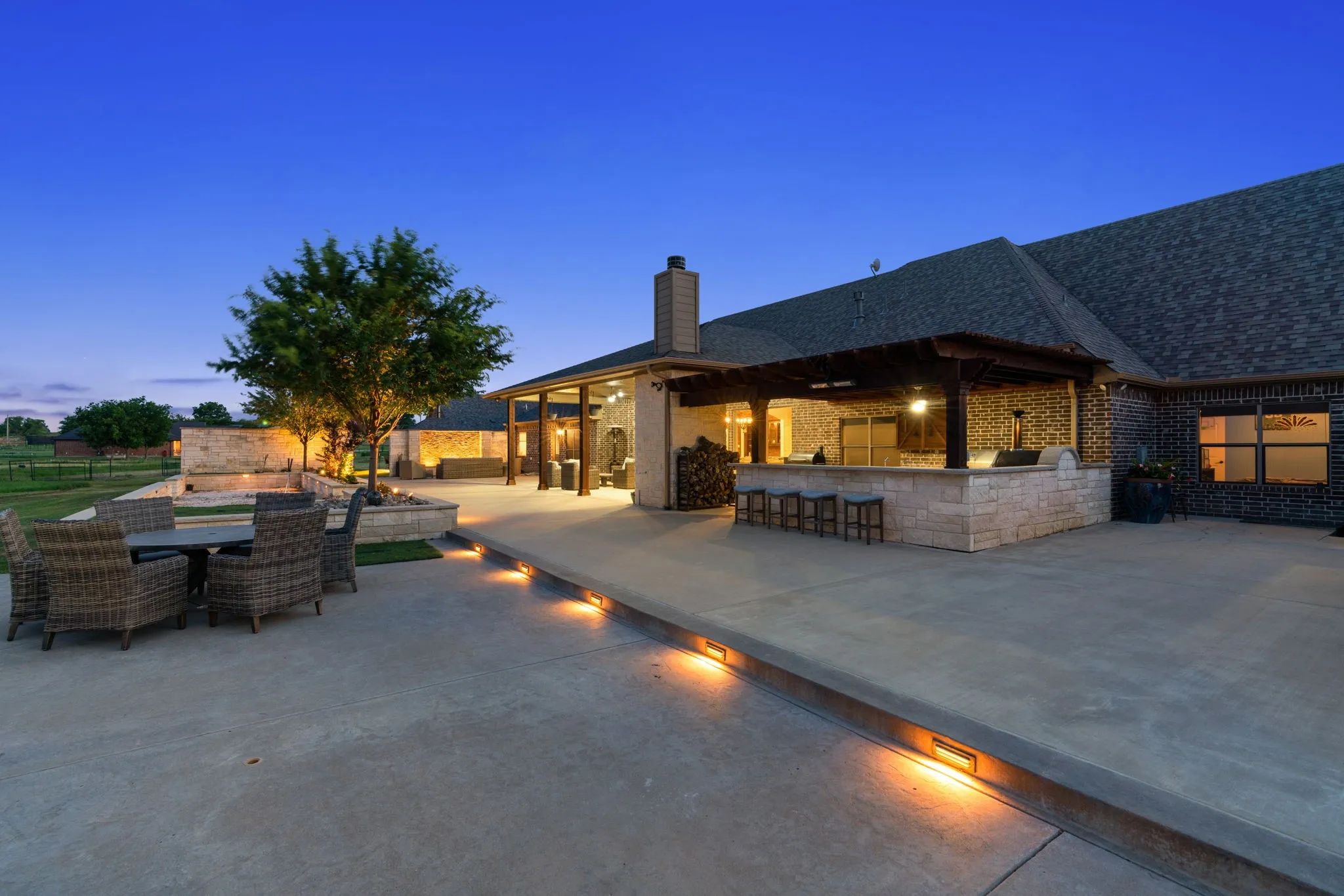 Expansive concrete patio with integrated lighting, a stone-clad outdoor kitchen with built-in appliances, a wooden pergola, and a stone-accented pool area