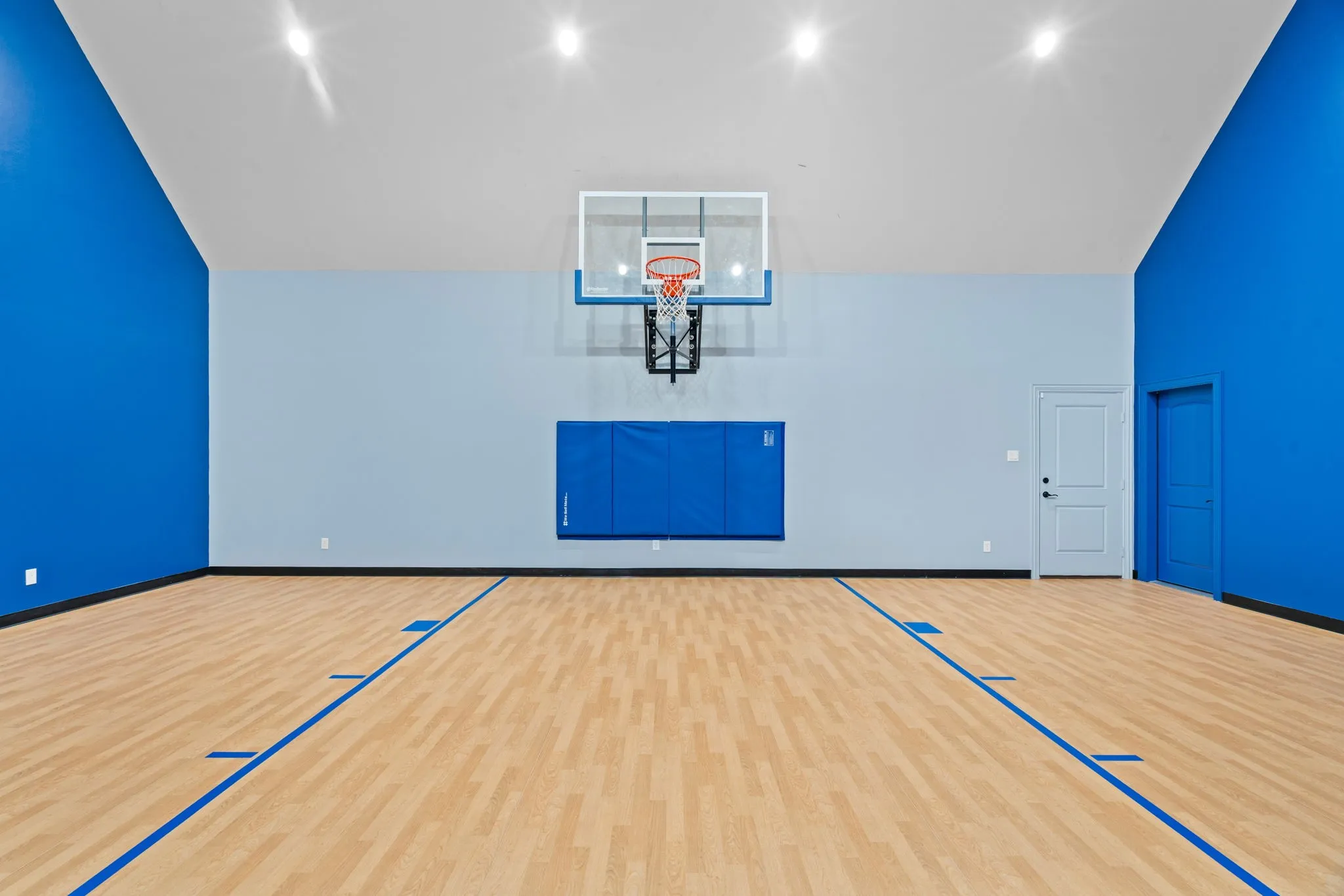 Indoor athletic court with wood-finish flooring, marked court lines, a basketball hoop, and padded wall protection
