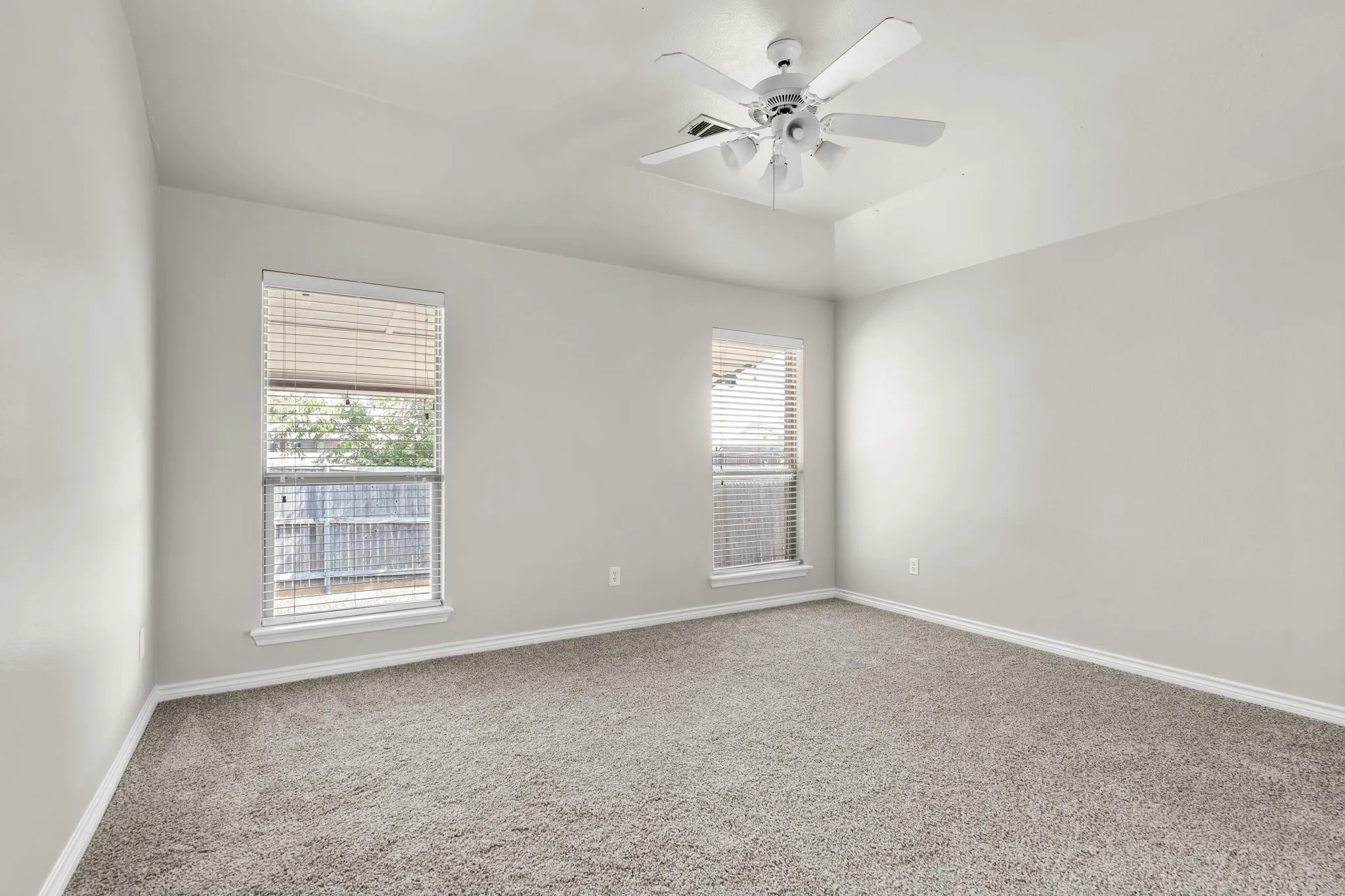 Neutral wall paint, plush carpet flooring, two windows with blinds, and a ceiling fan