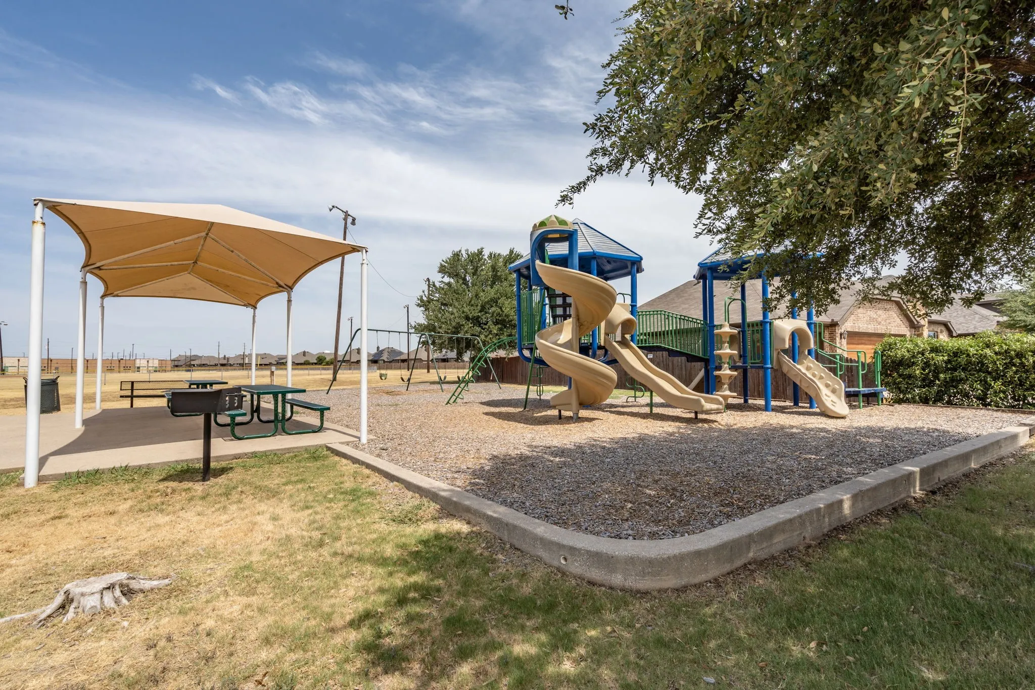 Community playground featuring multiple slides, swings, a shaded picnic area with a built-in grill, and a ground cover of wood chips