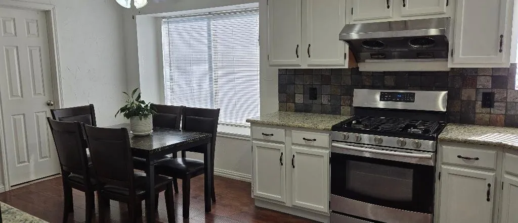 Wood-finish flooring throughout, white cabinetry with dark hardware, granite-style countertops, stainless steel range with matching hood, and a tile backsplash