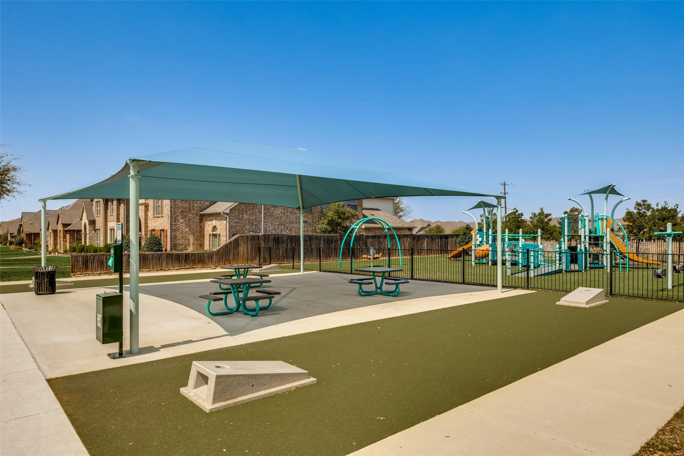 Outdoor recreational area featuring a shade structure, multiple picnic tables, a fenced playground with slides and climbing structures, and a turf ground surface