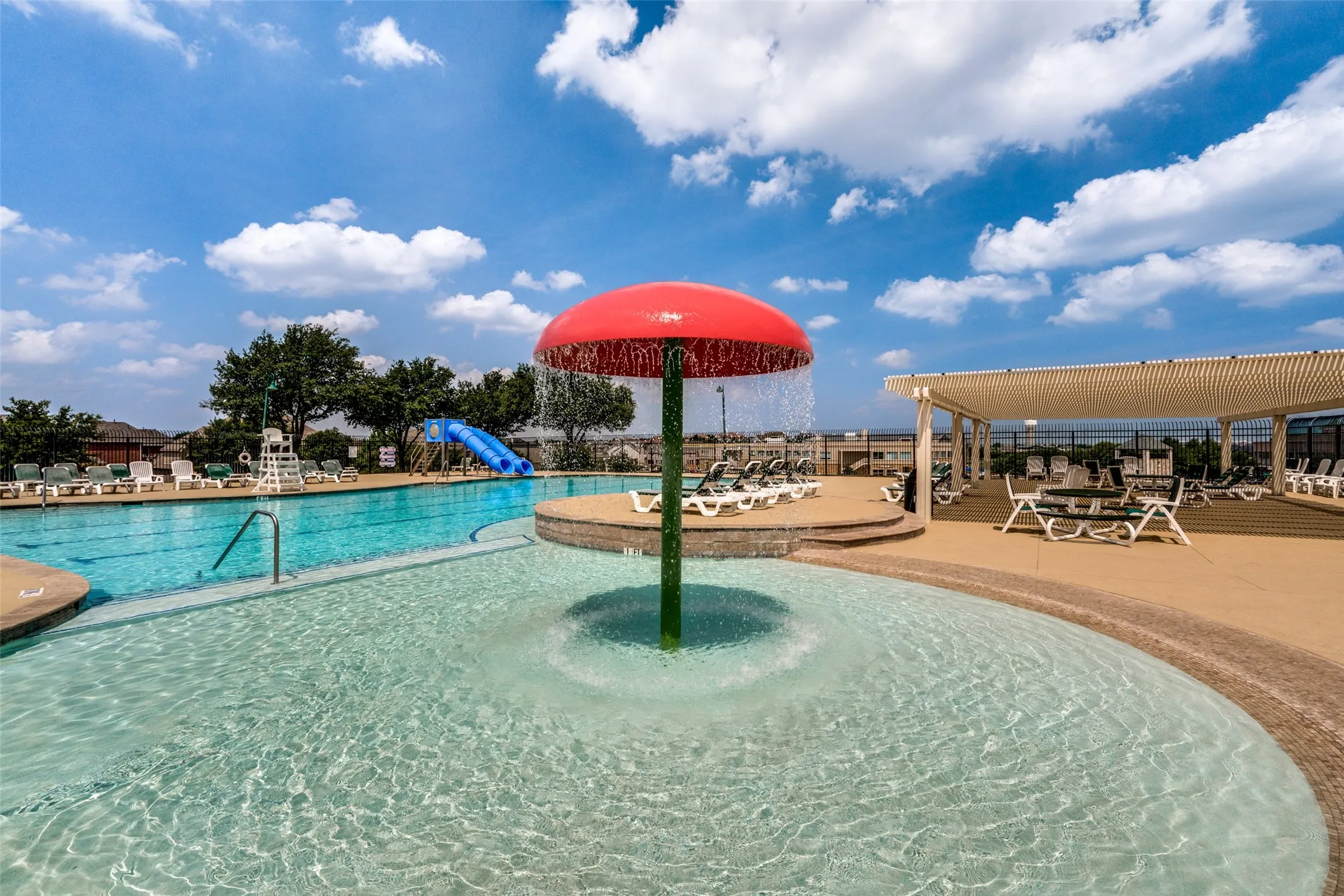 Resort-style pool area featuring a mushroom-shaped water feature, a main swimming pool with a slide, an expansive tanning deck, and a shaded pergola area