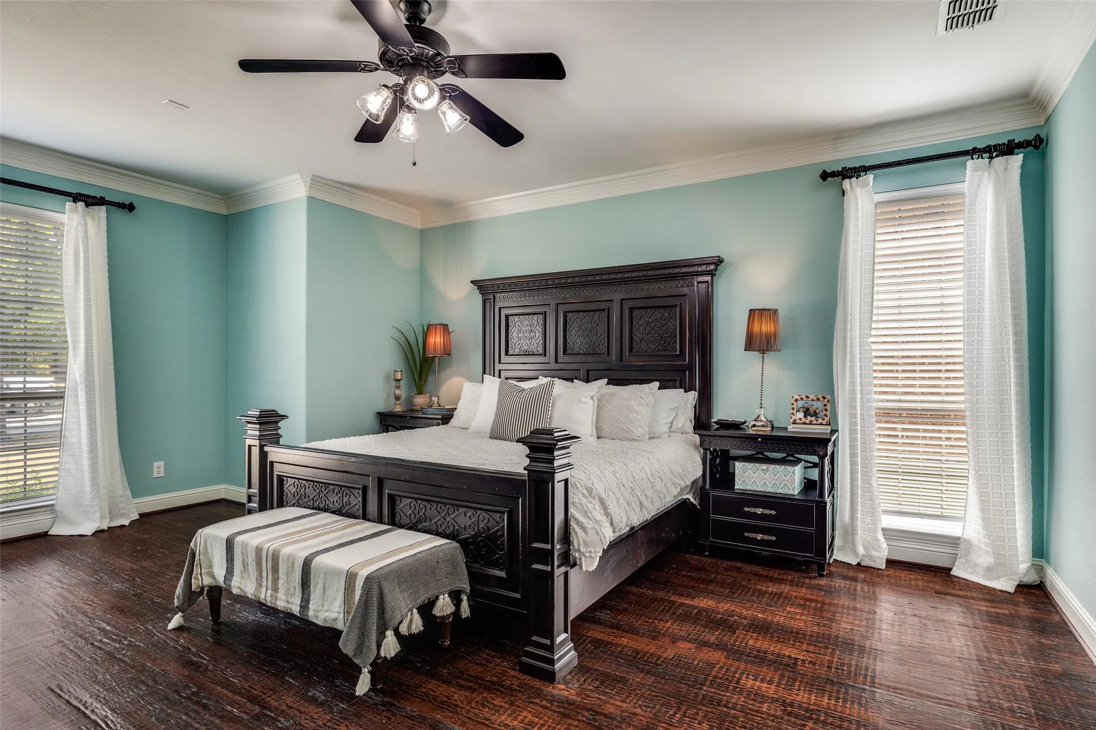 Master bedroom featuring turquoise walls, a dark wood four-poster bed frame, dark wood-finish flooring, ceiling fan with integrated lighting, dual glass doors with plantation shutters, and bedside nightstands with amber glass accent lamps
