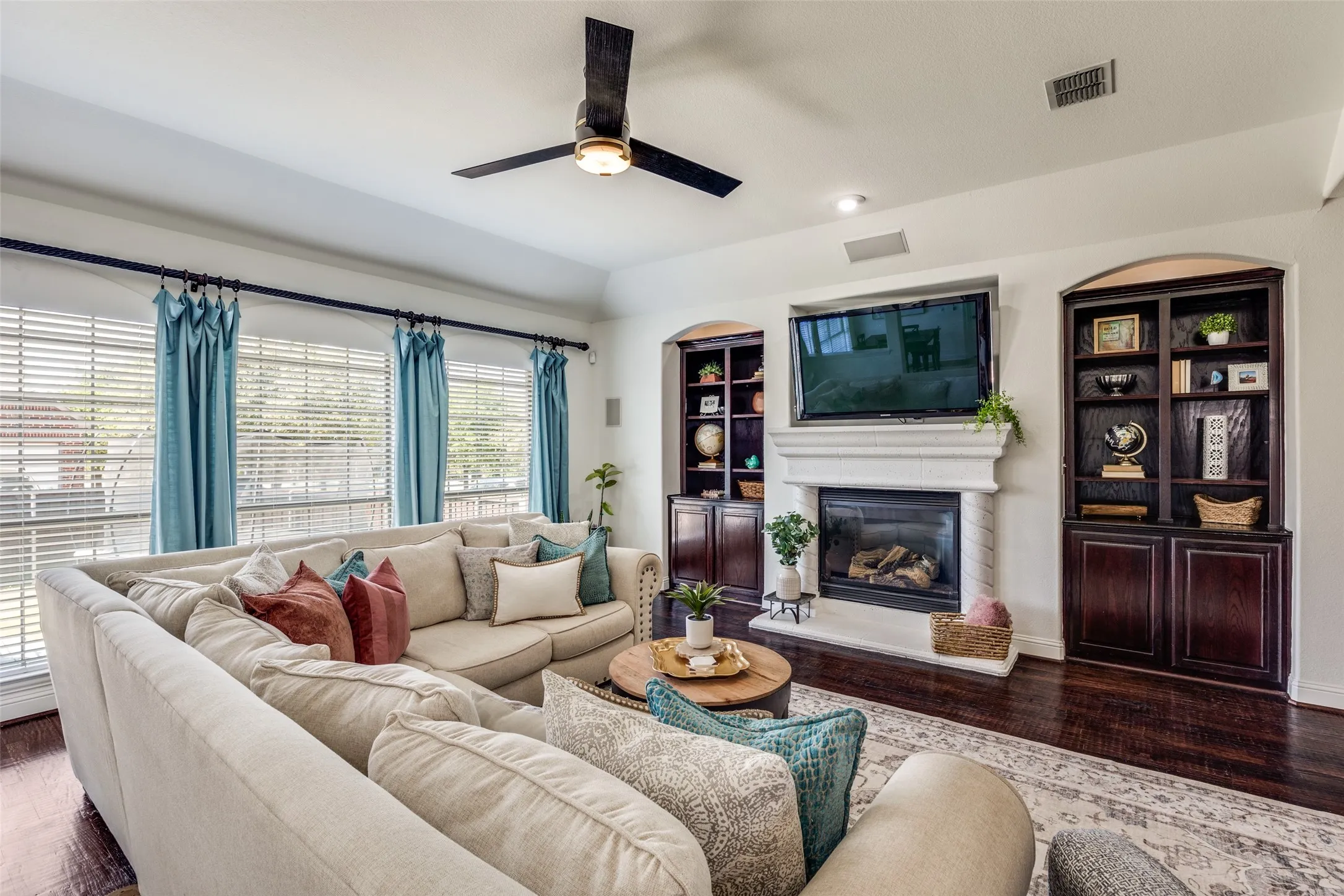 Tray ceiling with recessed lighting, integrated ceiling fan, warm wood-finish flooring, built-in display cabinetry, and a fireplace with a mantel