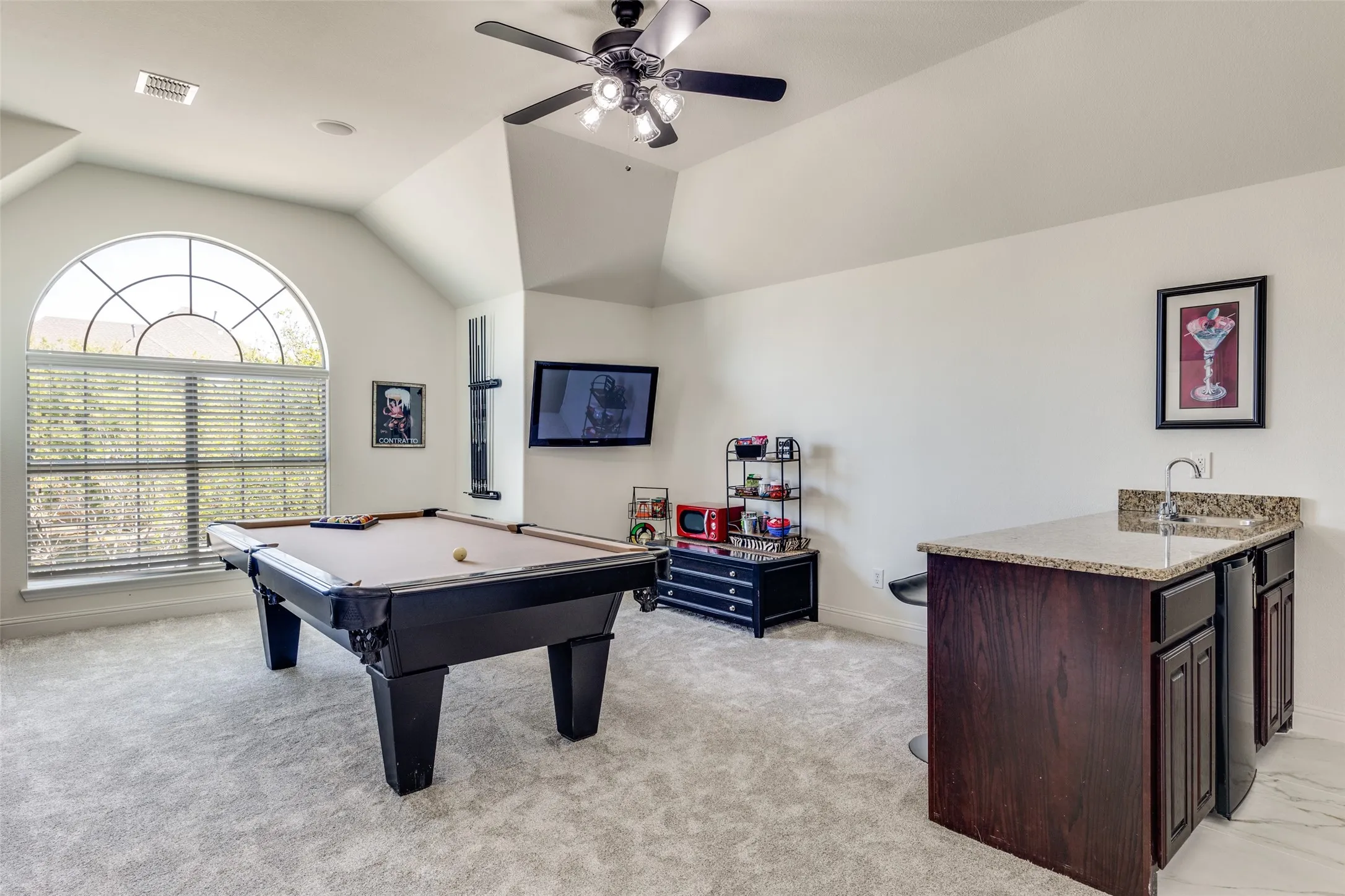 Arched window with blinds, ceiling fan with light fixture, built-in wet bar with granite-style countertop, and dark wood-finish cabinetry