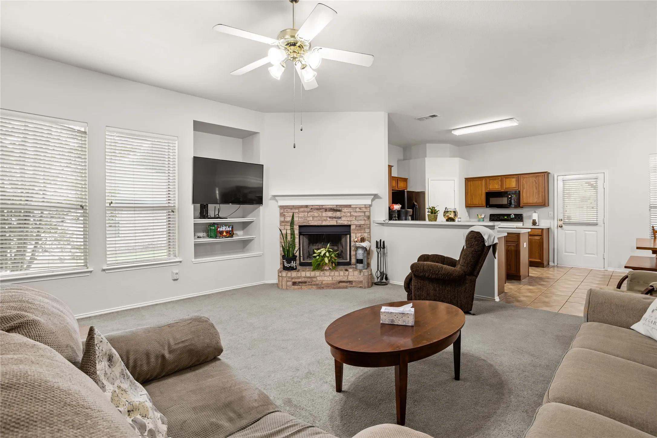 Spacious living area featuring a brick fireplace, built-in shelving, carpet flooring, and a ceiling fan