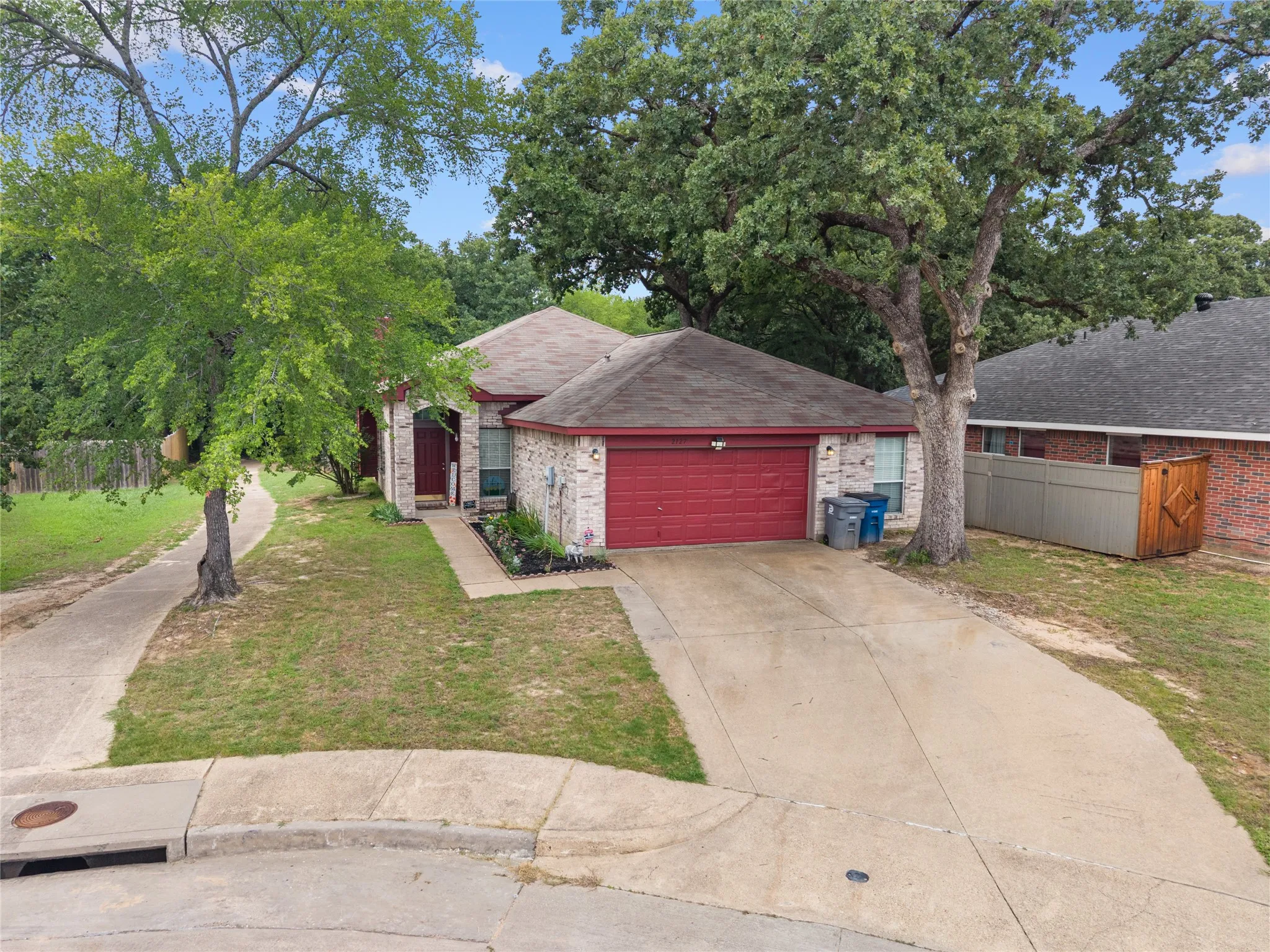 Charming one-story brick home featuring a red garage door, mature landscaping with established trees, and a concrete driveway