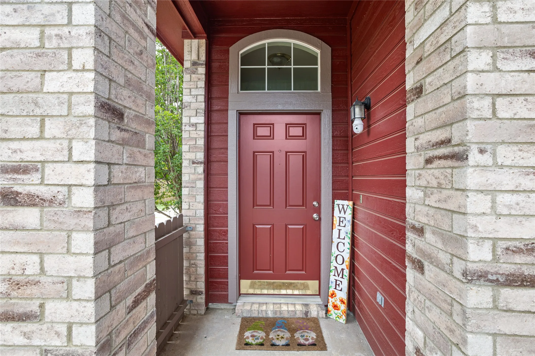 Red paneled entry door with an arched transom window, flanked by light-toned brick columns and red horizontal siding