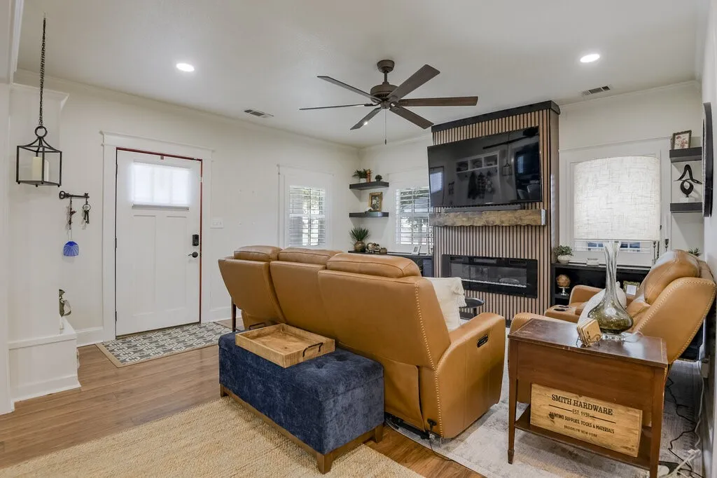 Welcoming living room featuring warm wood-finish flooring, a ceiling fan with integrated lighting, and a fireplace with wood surround and shelving