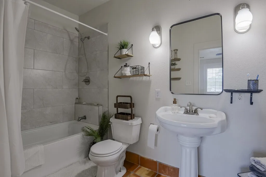Bright bathroom featuring a pedestal sink with chrome fixtures, subway tile shower surround with glass enclosure, toilet with tank, wood-finish flooring, framed mirror with integrated lighting, and open shelving with decorative baskets for storage organization