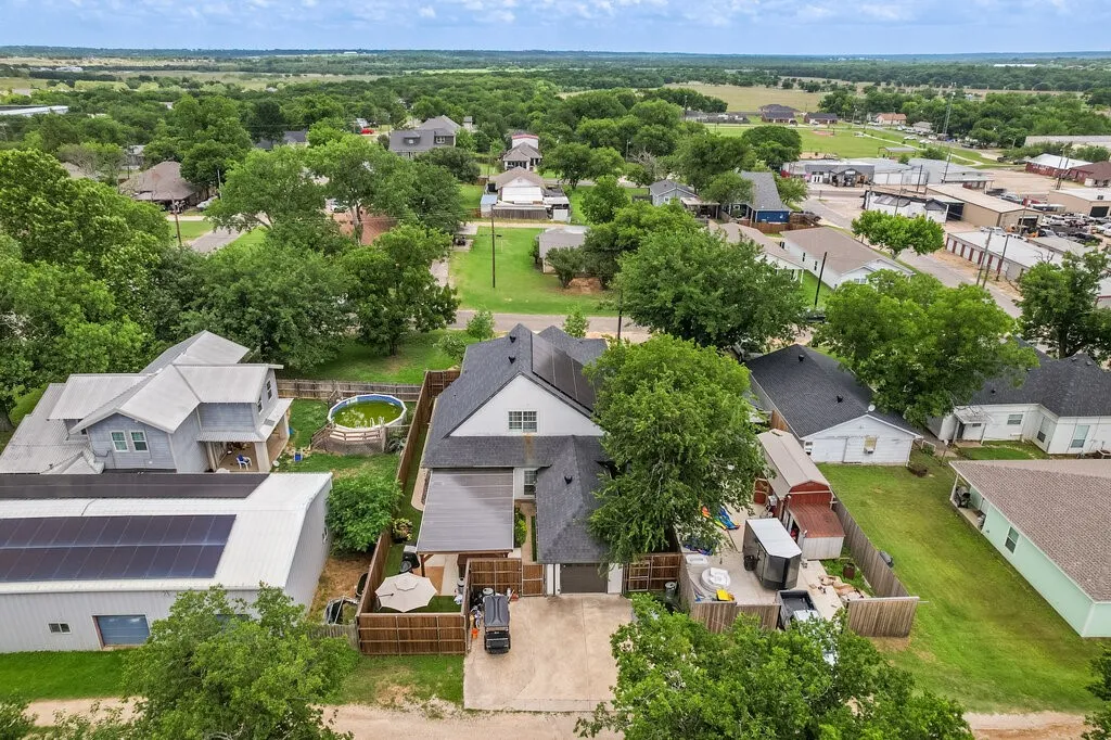Aerial view showcasing a contemporary home with dark roof and light exterior, situated on a spacious lot surrounded by mature landscaping