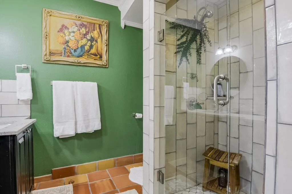 Spa-inspired ensuite bathroom featuring vibrant emerald green accent walls, terra cotta tile flooring, white subway tile shower surround with frameless glass enclosure, polished chrome fixtures, and decorative wall-mounted shelving