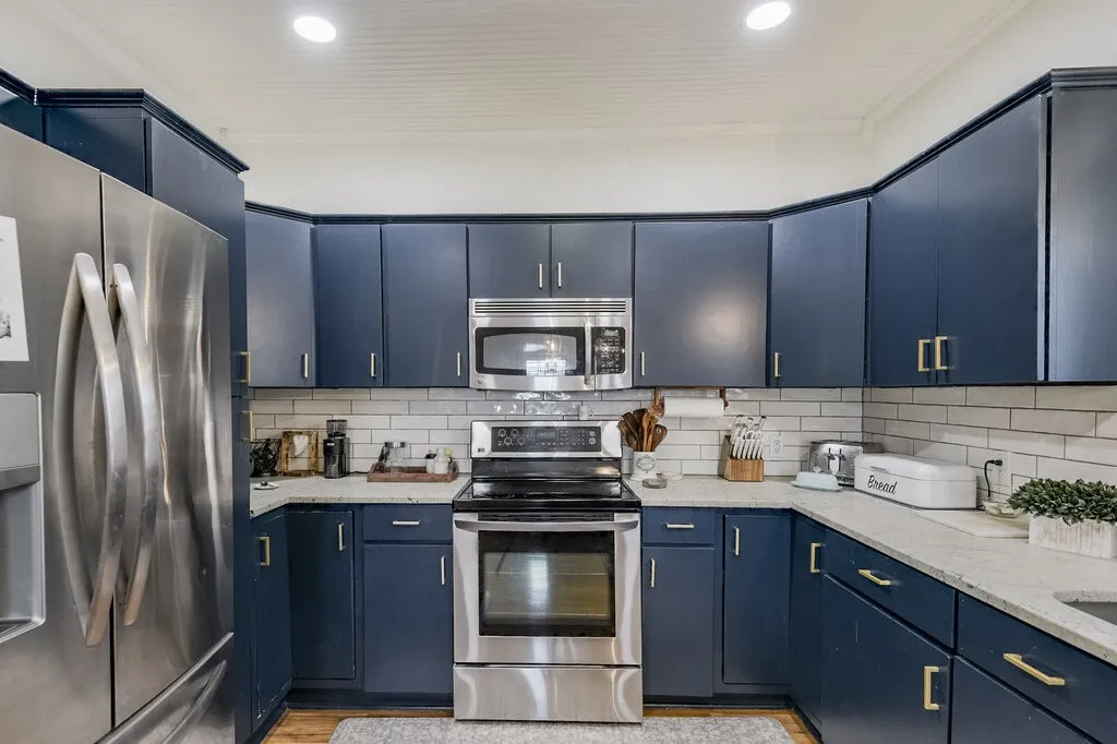 Modern kitchen featuring custom navy cabinetry with brushed gold hardware, stainless steel appliances including a built-in microwave and gas range, quartz countertops, subway tile backsplash, and recessed ceiling lighting