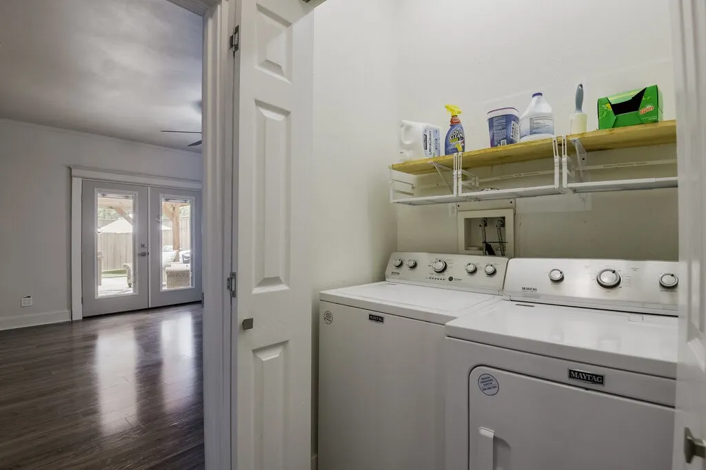 Dedicated laundry room featuring side-by-side washer and dryer with stainless steel finishes, open shelving with storage containers, and direct access to the main living spaces through a white six-panel door