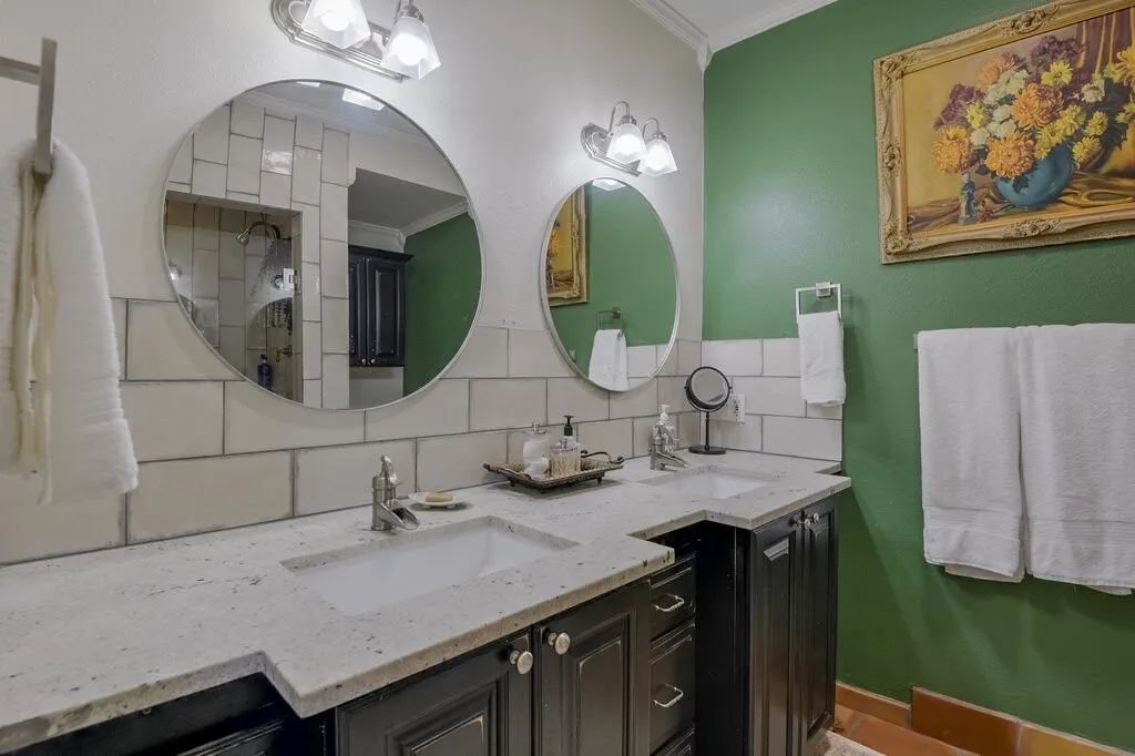 Spa-inspired primary bathroom featuring dual circular mirrors with integrated lighting, marble-topped vanity with black cabinetry, white subway tile backsplash, polished nickel fixtures, and a rich jewel-tone accent wall complemented by coordinating artwork and soft-close towel warming hardware