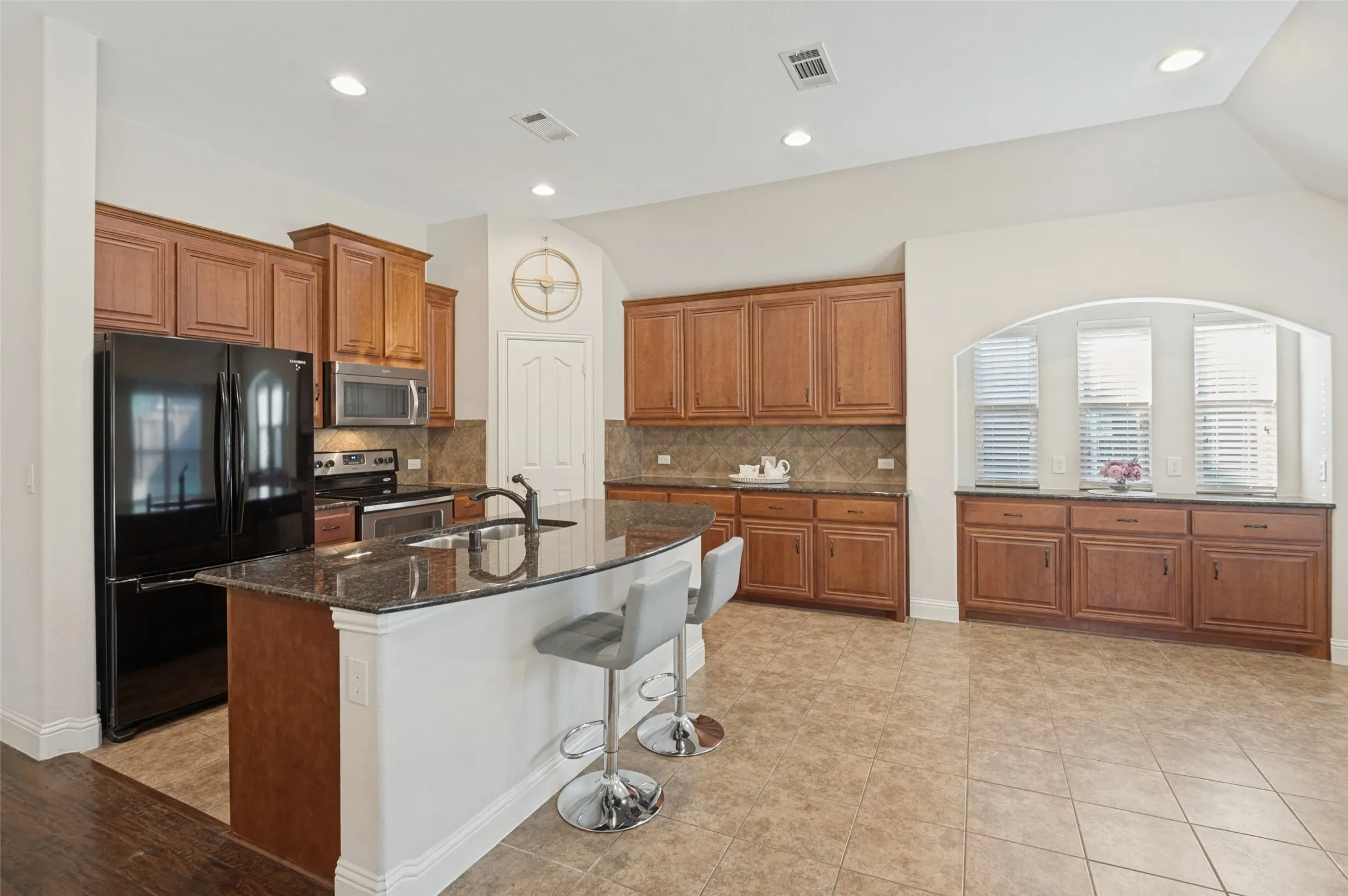 Kitchen with wood cabinetry, black appliances, a central island with a sink, and tile flooring