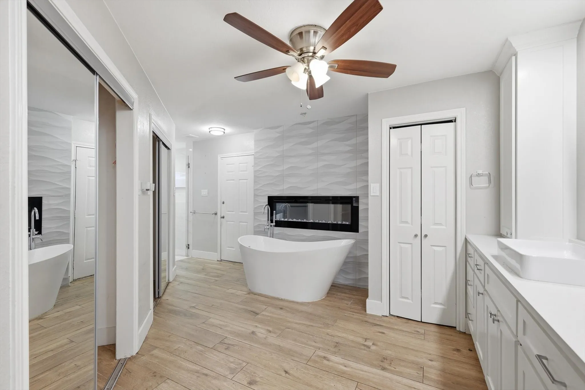 Wood-finish flooring, freestanding soaking tub, wall-mounted electric fireplace, white floating vanity with vessel sink, and a ceiling fan