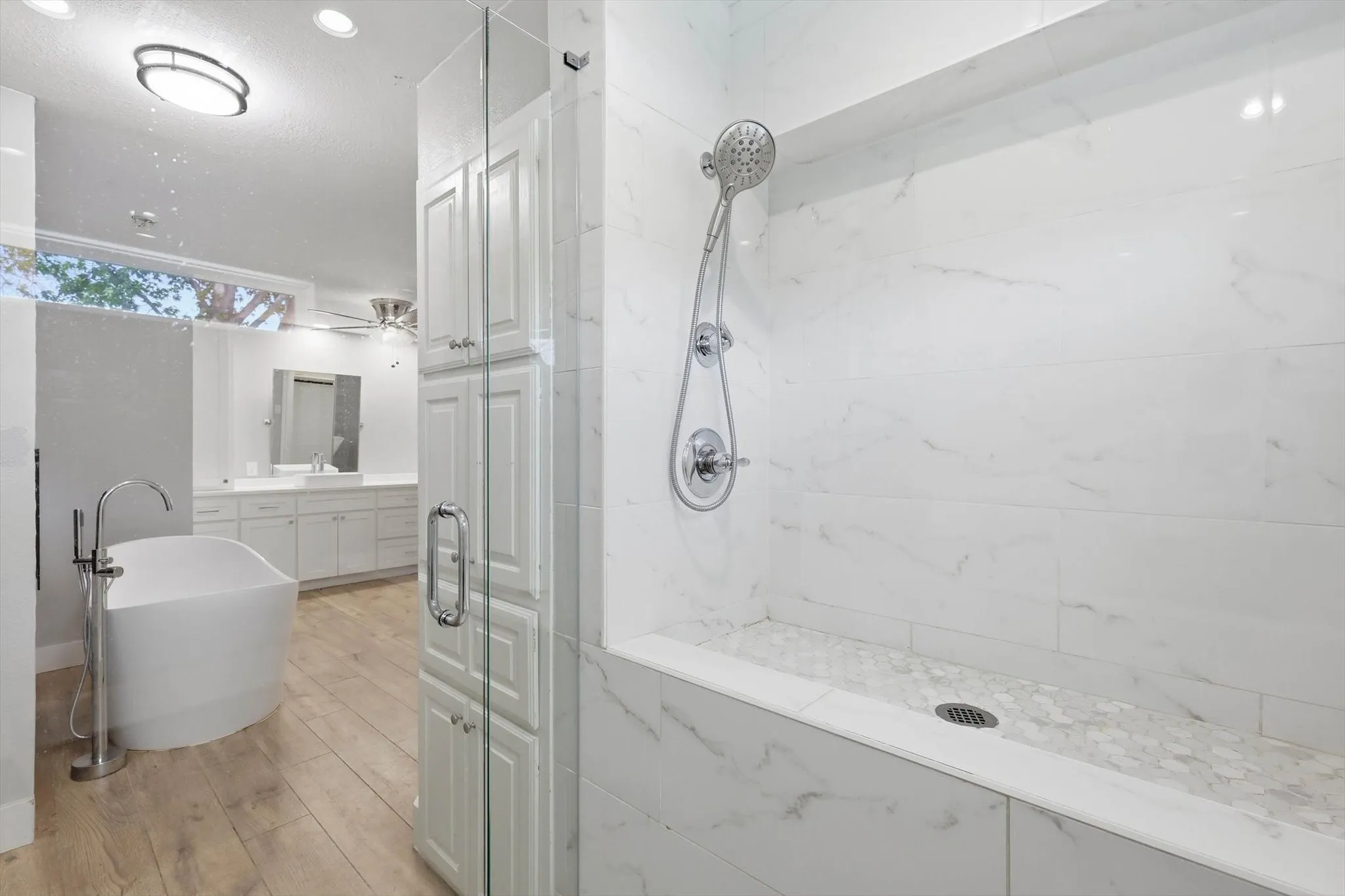 Walk-in shower with marble-style tile walls and mosaic floor, freestanding soaking tub with floor-mounted faucet, wood-finish flooring, and white cabinetry