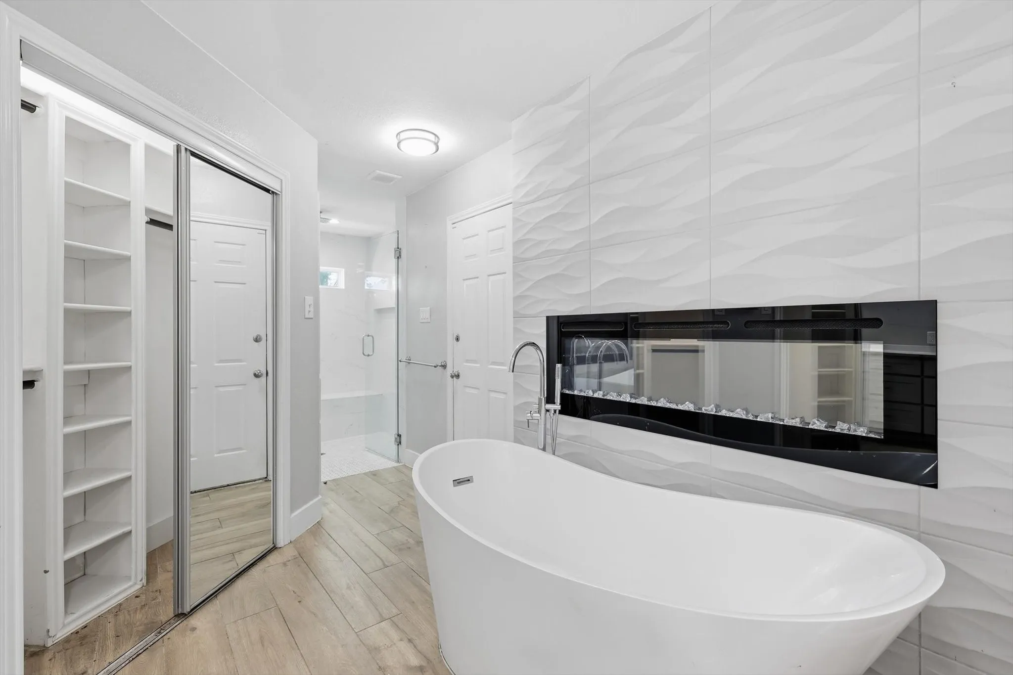Freestanding soaking tub with a floor-mounted faucet, electric fireplace with a textured tile surround, wood-finish flooring, and a walk-in shower with a glass enclosure