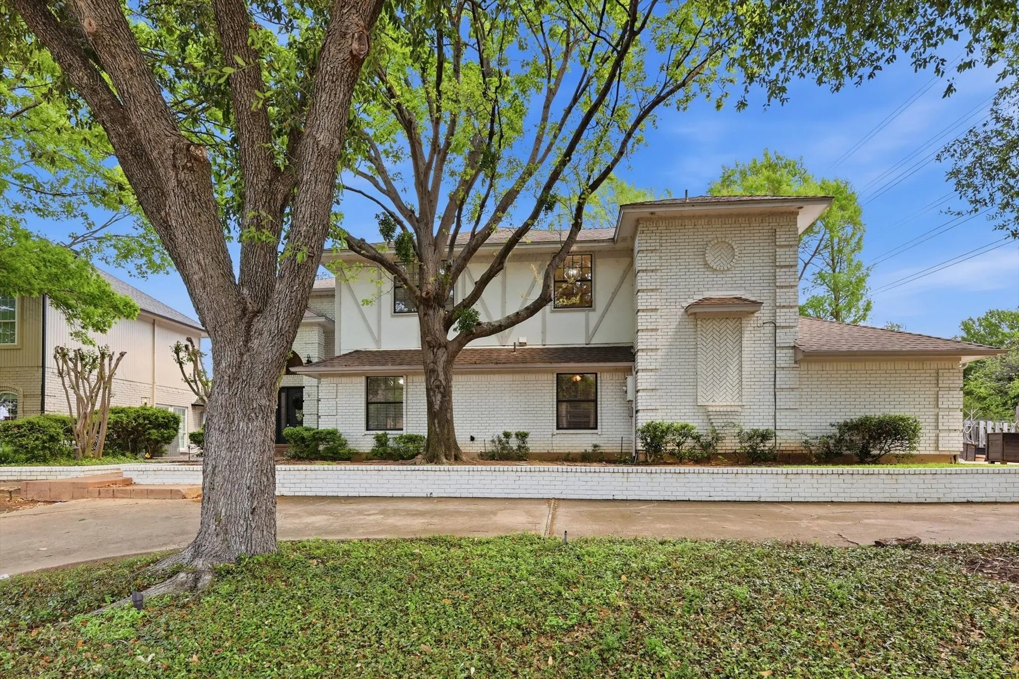 Stucco and brick exterior with contrasting trim, mature trees, manicured landscaping, and a low brick wall