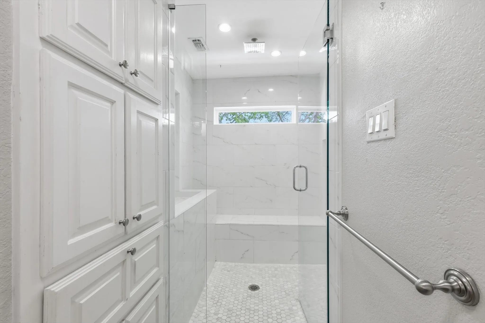 Walk-in shower with clear glass enclosure, a built-in bench, and white hexagonal floor tiling